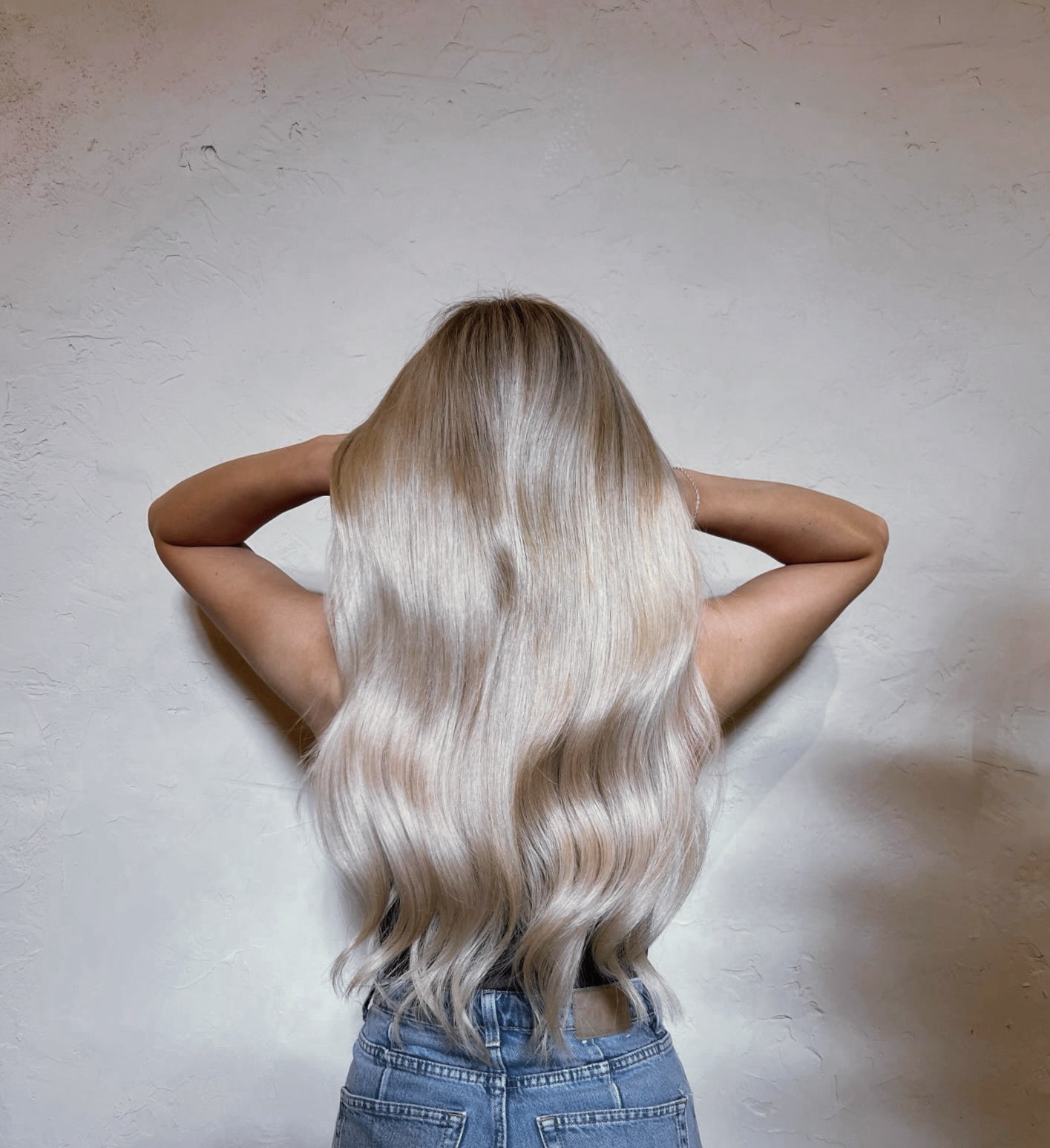Person with long, wavy blonde hair facing away, arms raised, wearing a blue top, against a plain wall.