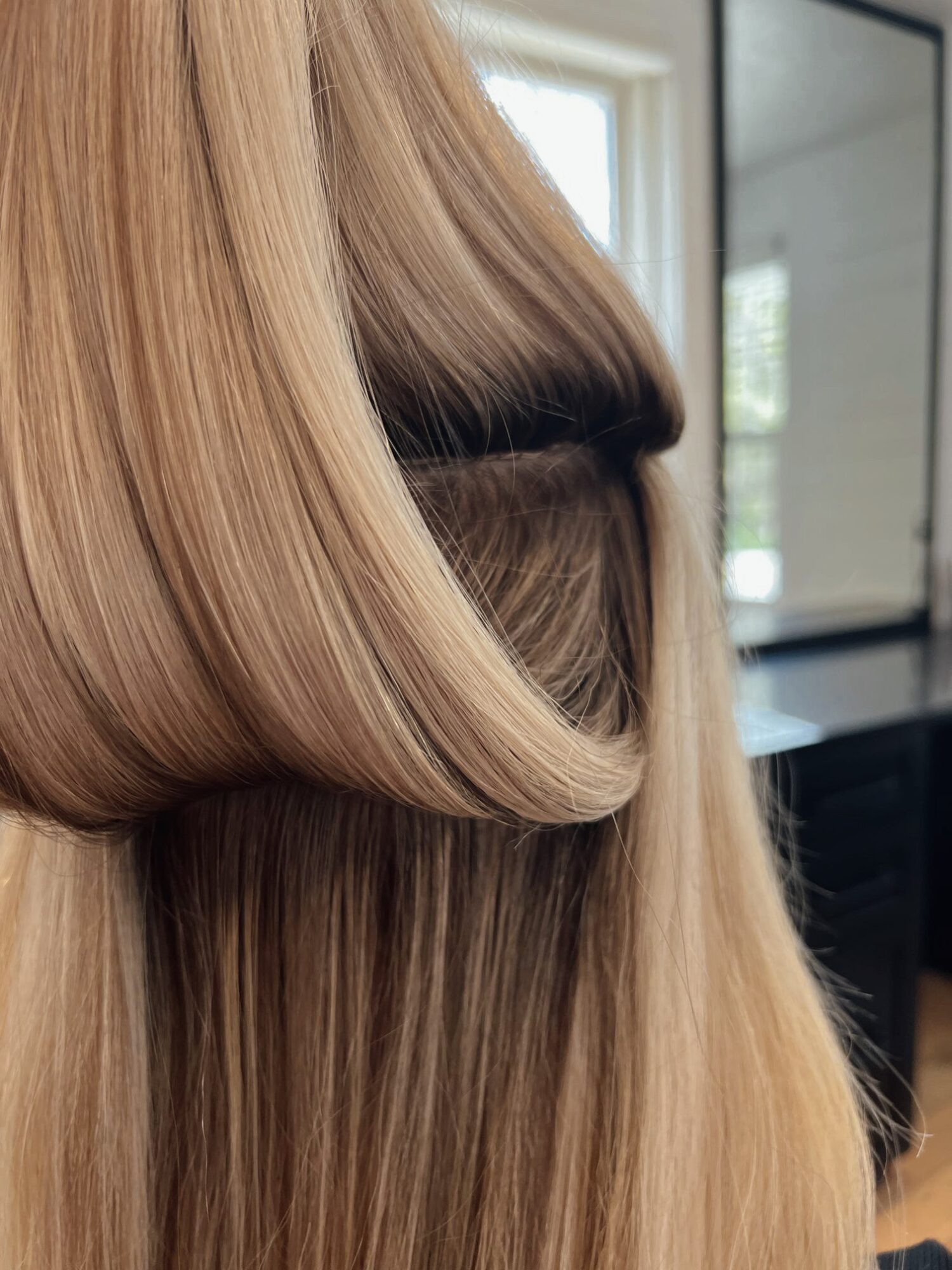 Close-up of blonde hair with a section lifted, showing smooth, shiny strands and a mirror in the background.