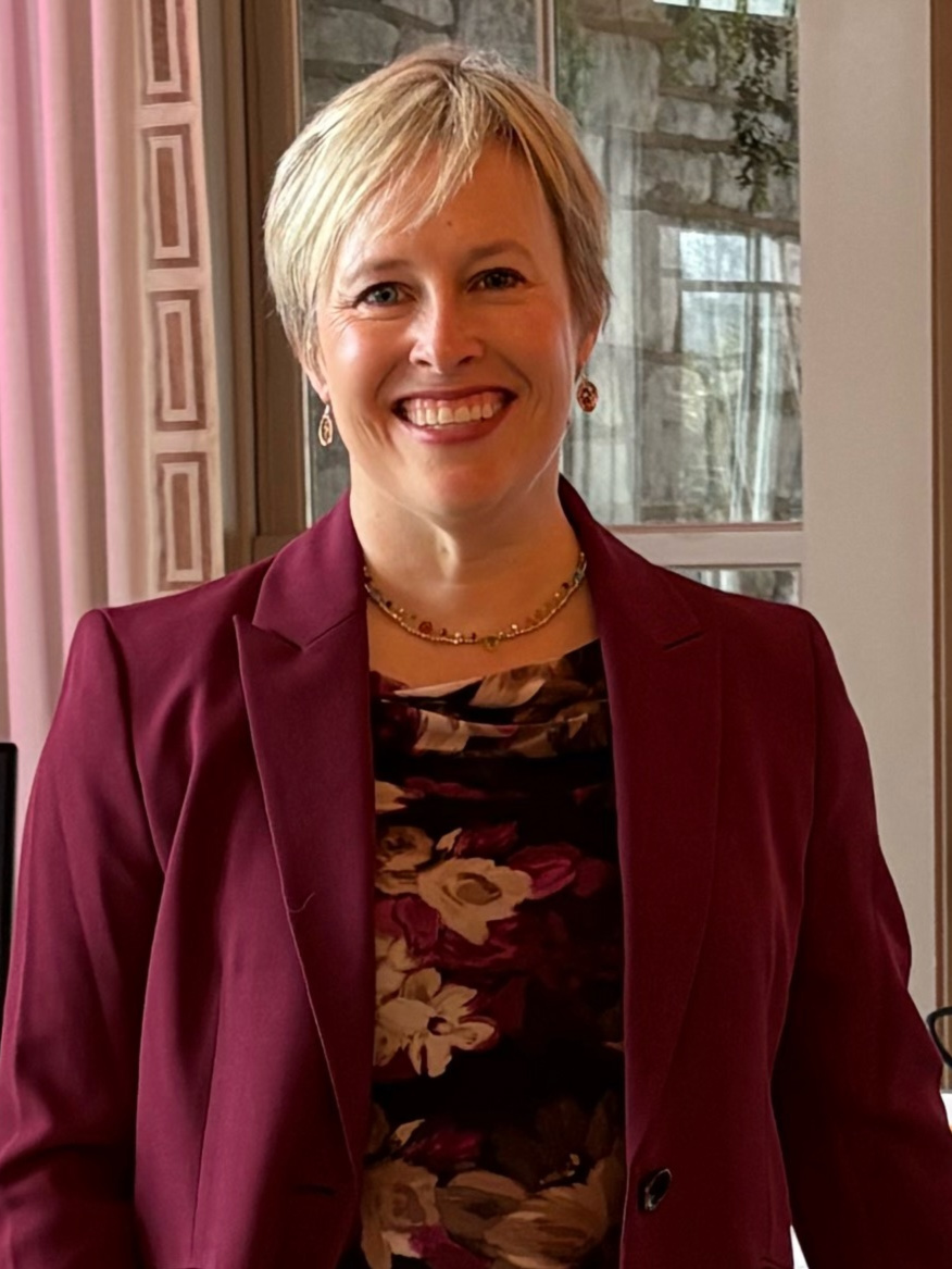 Woman with short blonde hair smiling, wearing a maroon blazer and floral top, standing indoors near a window.