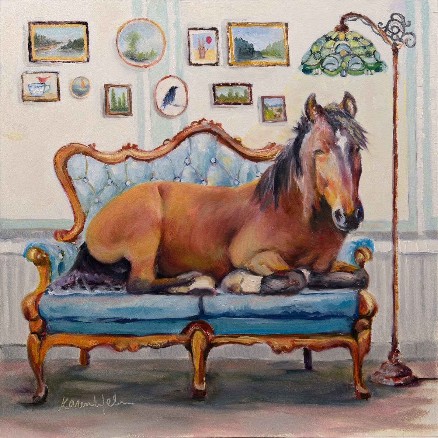 A horse lying on a vintage blue sofa in a room with framed pictures on the wall and a green lamp. Word count: 20