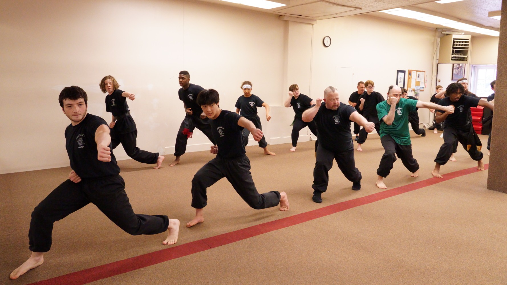 Group of people practicing martial arts in a room, performing a synchronized move with arms extended and legs bent.