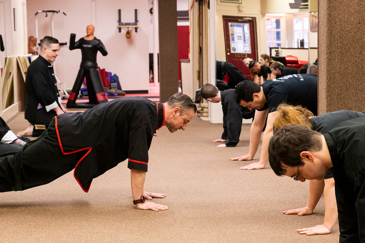 People practicing yoga or stretching on the floor in a room with large windows and a mirror, led by an instructor.