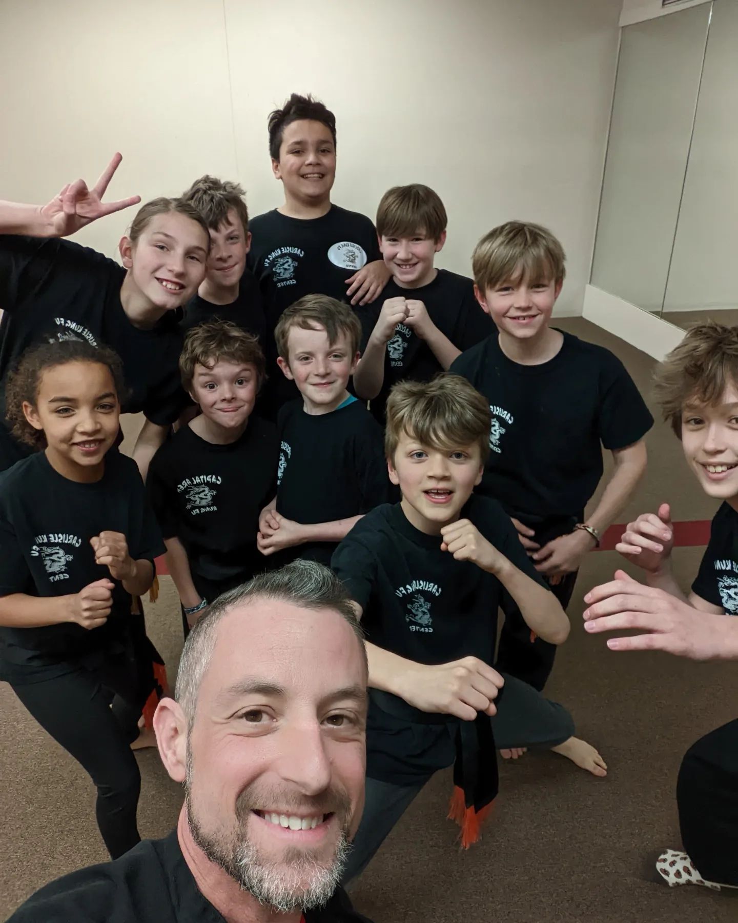 Group of children and an adult man smiling indoors, some making peace signs, wearing matching black shirts.