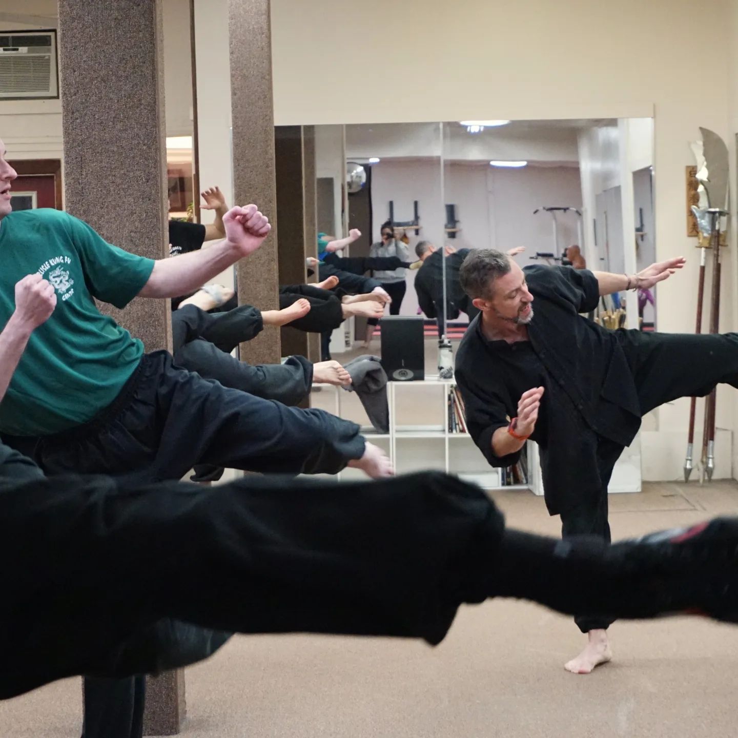 Instructor leading a group in a martial arts class, performing side kicks in a studio with mirrors and equipment.