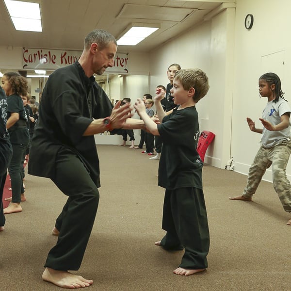 A martial arts instructor and a young student practicing in a training room with other students in the background.