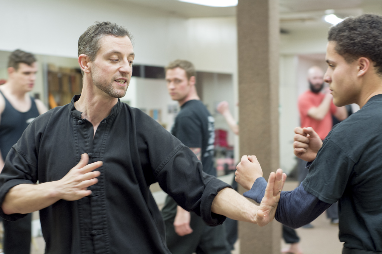 Two men practicing martial arts in a gym, one with a black uniform and the other with a dark shirt, engaging in a training exercise.