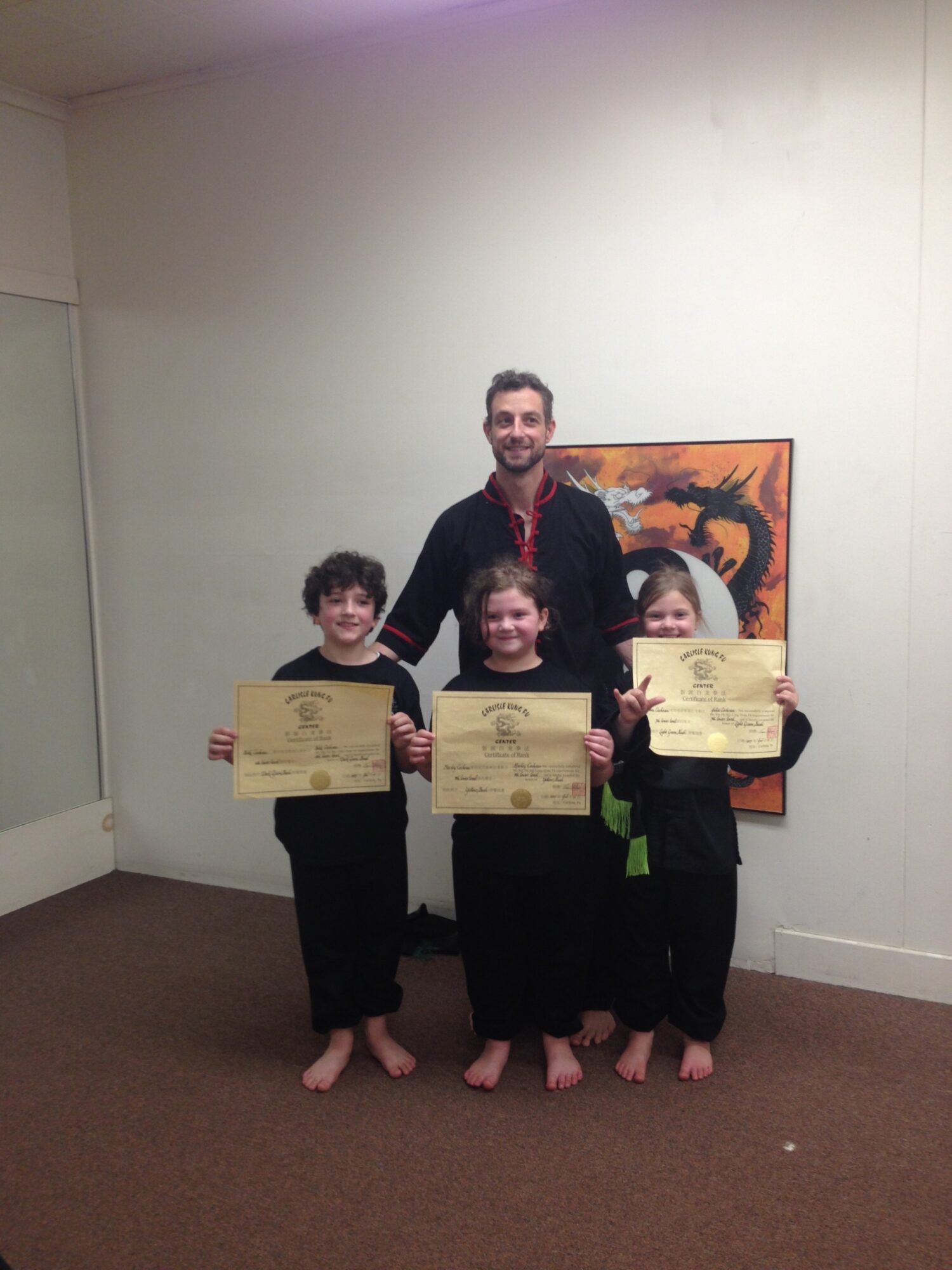 Four people, three children and one adult, standing indoors holding certificates, with a colorful painting on the wall behind them.