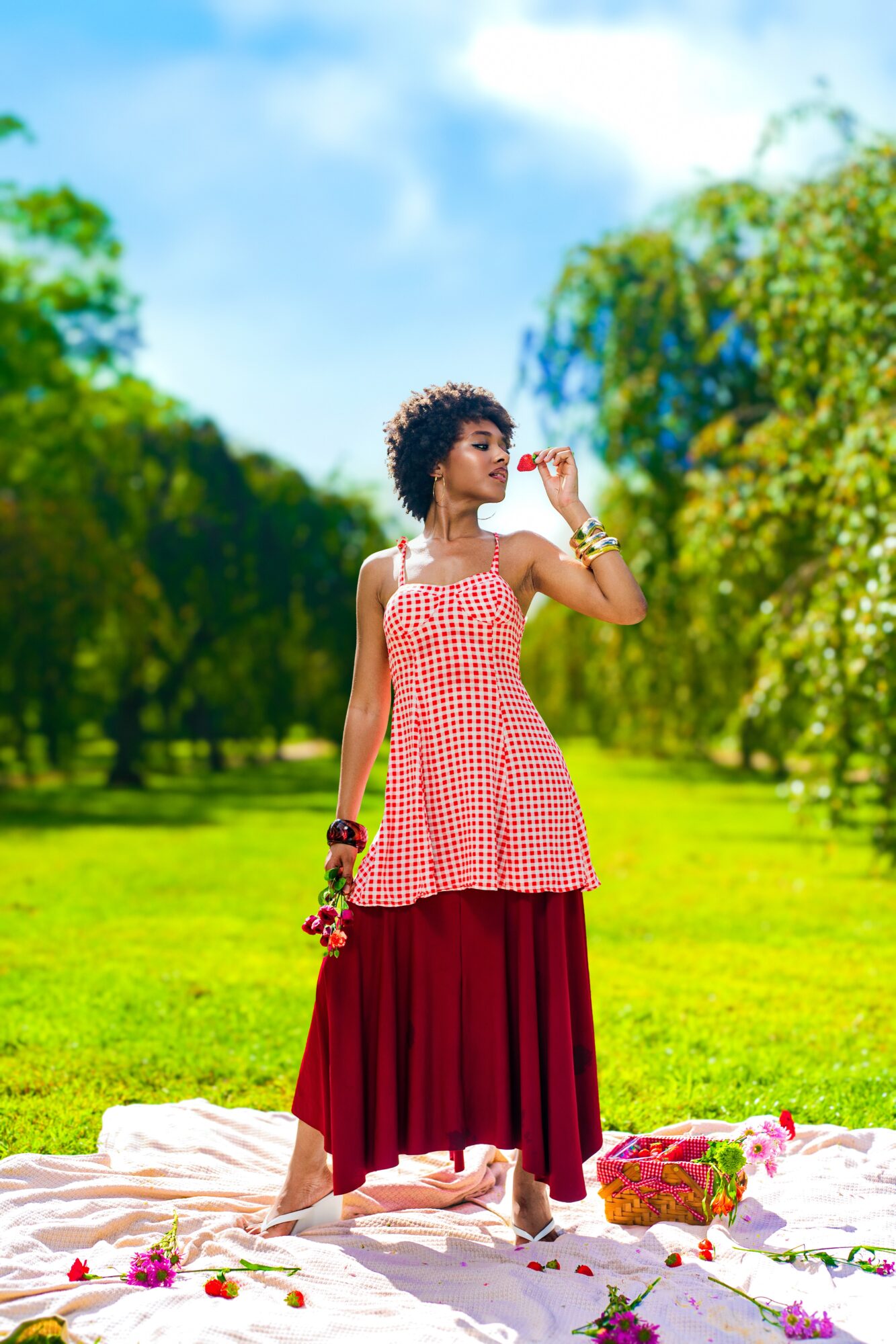 Woman with curly hair in a red and white checkered top and long red skirt stands outdoors on grass, holding a bracelet, with trees and blue sky in background.