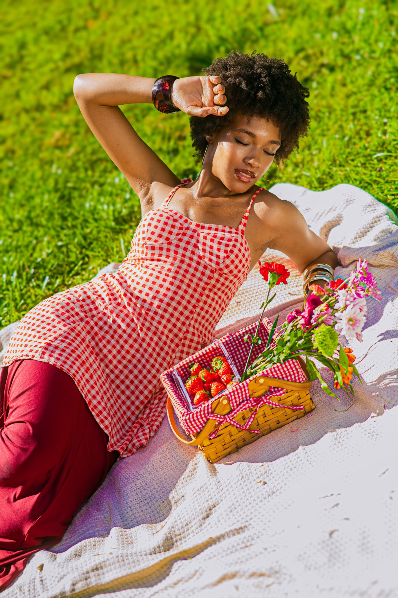 Young woman with curly hair lying on grass, wearing a red and white checkered dress, surrounded by flowers and a basket of strawberries.