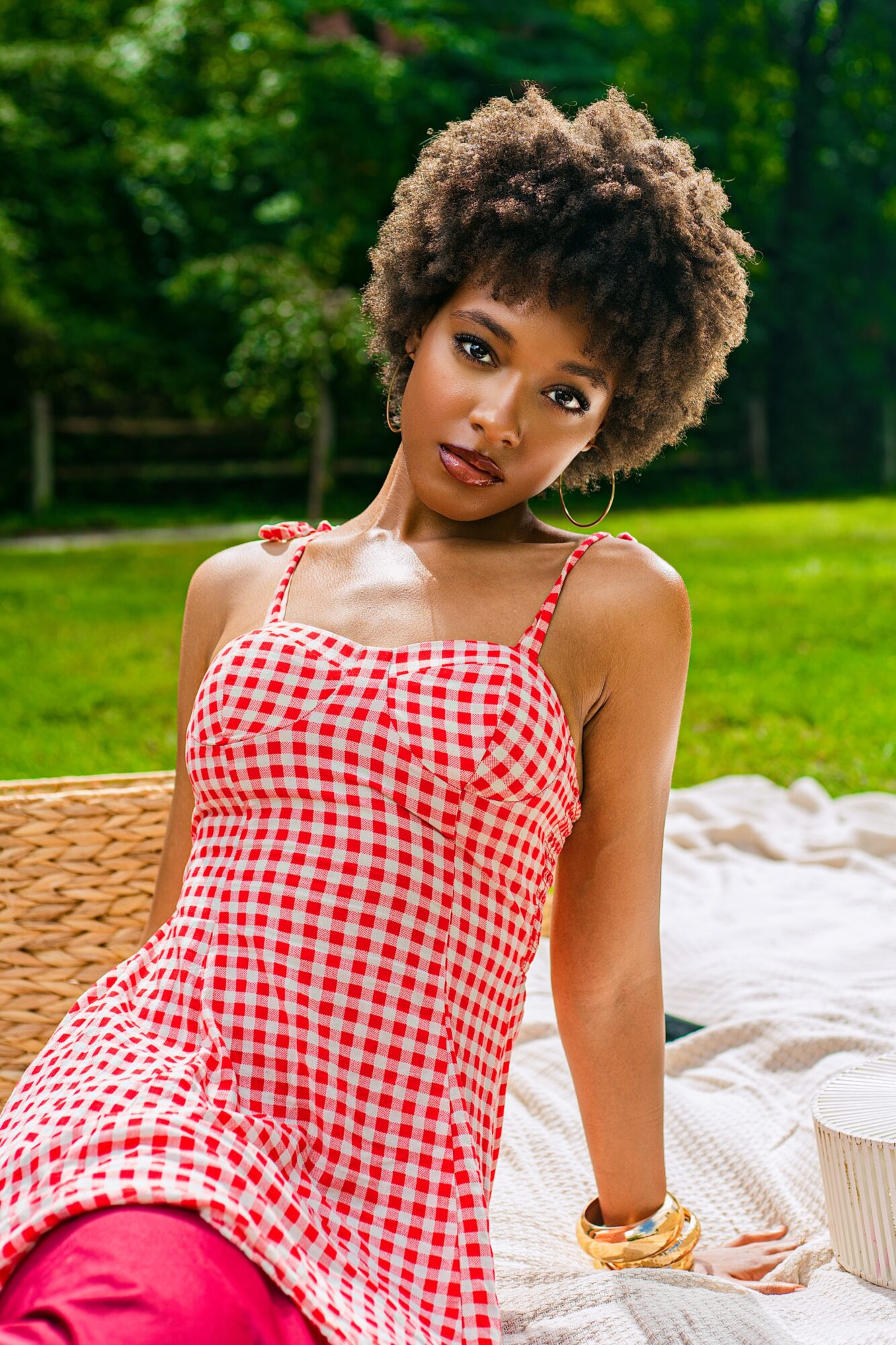 Young woman with curly hair in a red and white checkered dress outdoors, sitting on a wicker chair, green background.