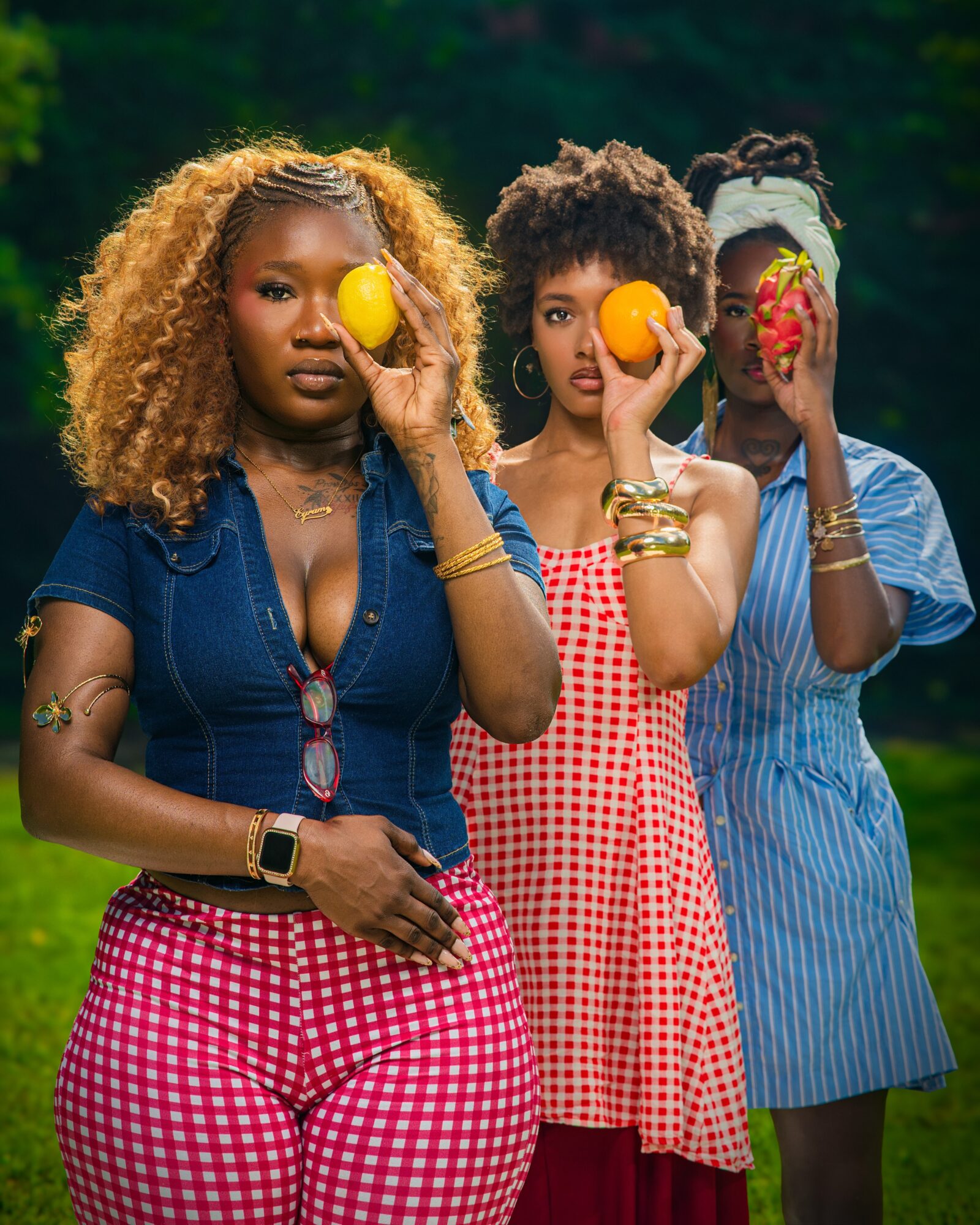 Three women outdoors holding fruits in front of their faces, with trees in the background.