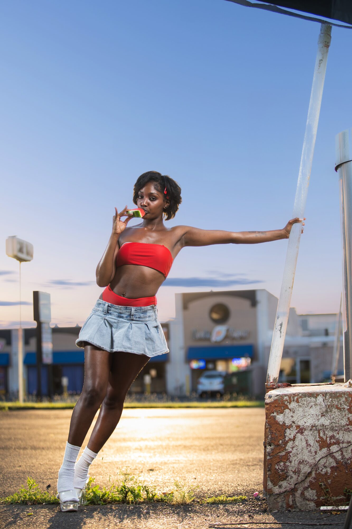 Young woman in red top and denim skirt leaning against a pole outdoors, with a clear blue sky and buildings in background.