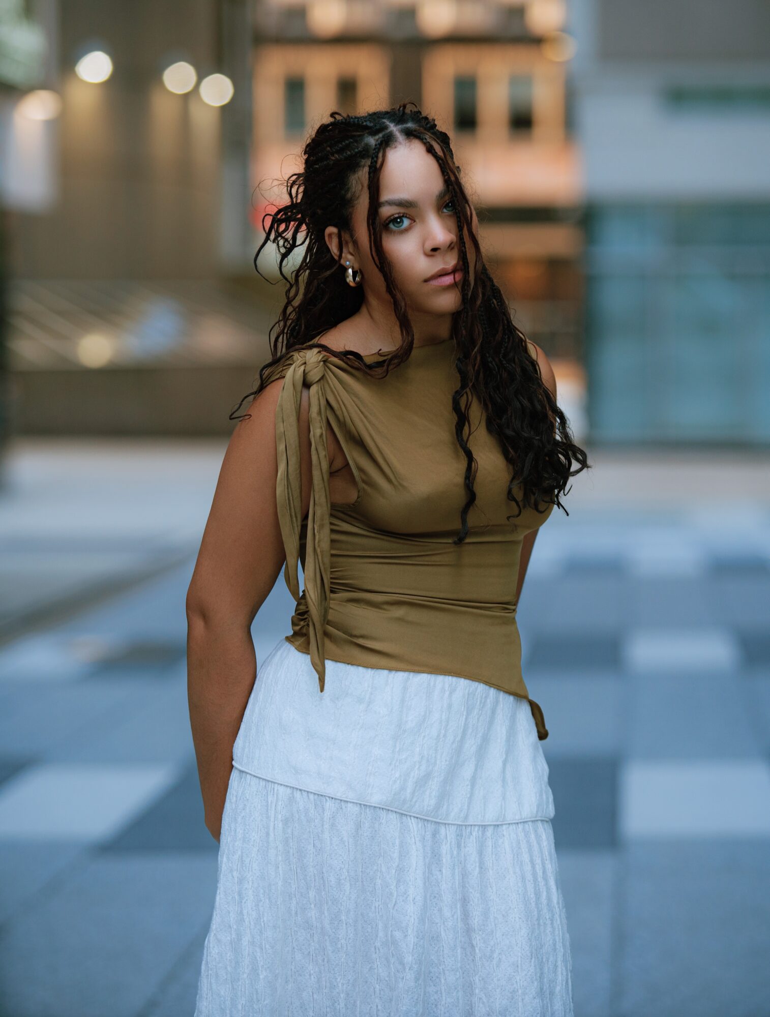 Young woman with long curly hair stands outdoors in an urban setting, wearing a brown top and white skirt.