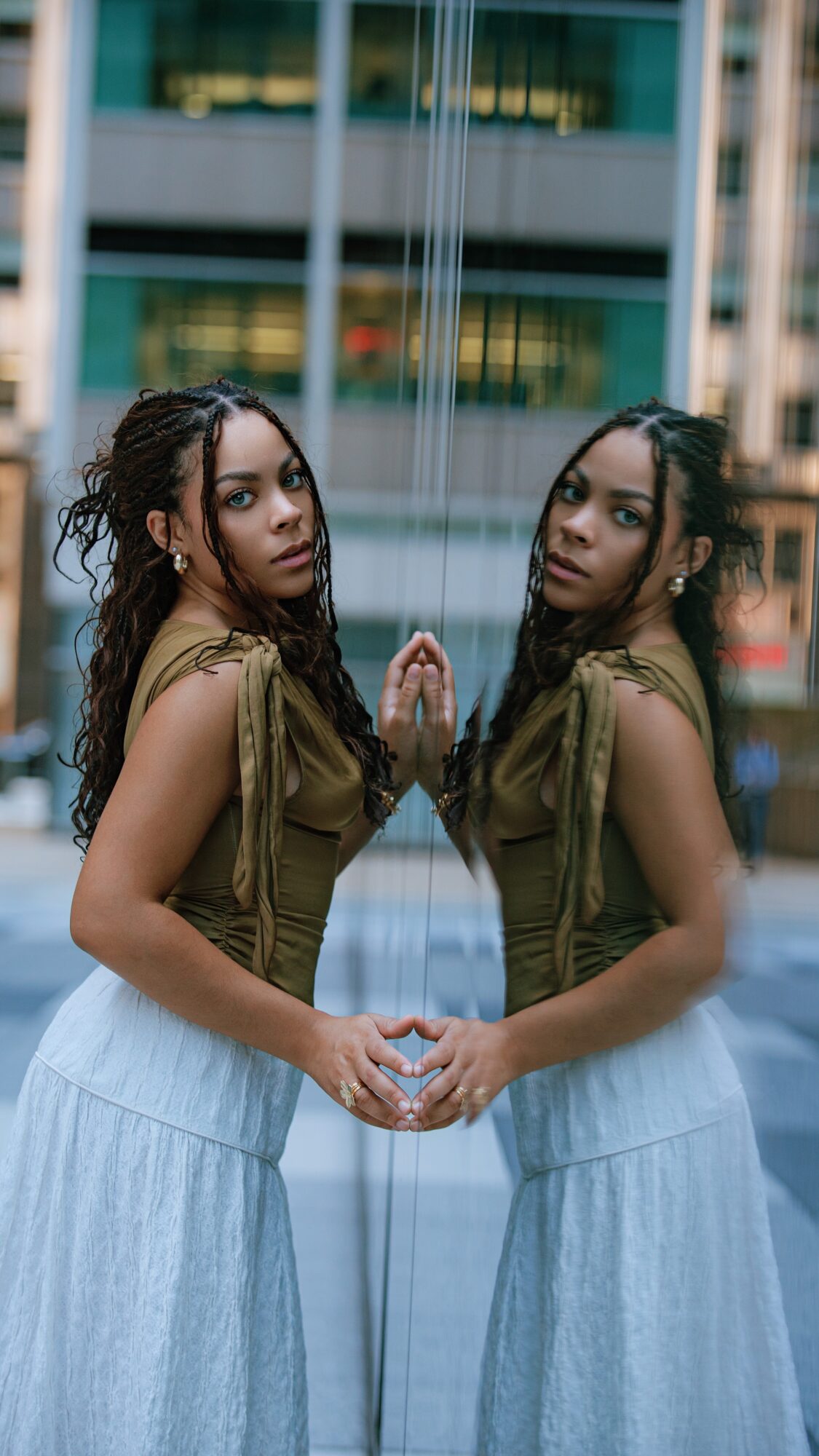 Two women stand facing each other with hands touching, in front of a reflective surface and modern building. 20 words.
