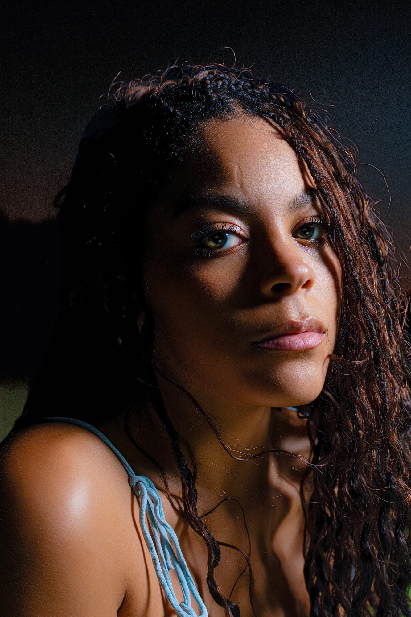 Close-up of a woman with curly hair and intense gaze, dark background, lighting highlights her face.