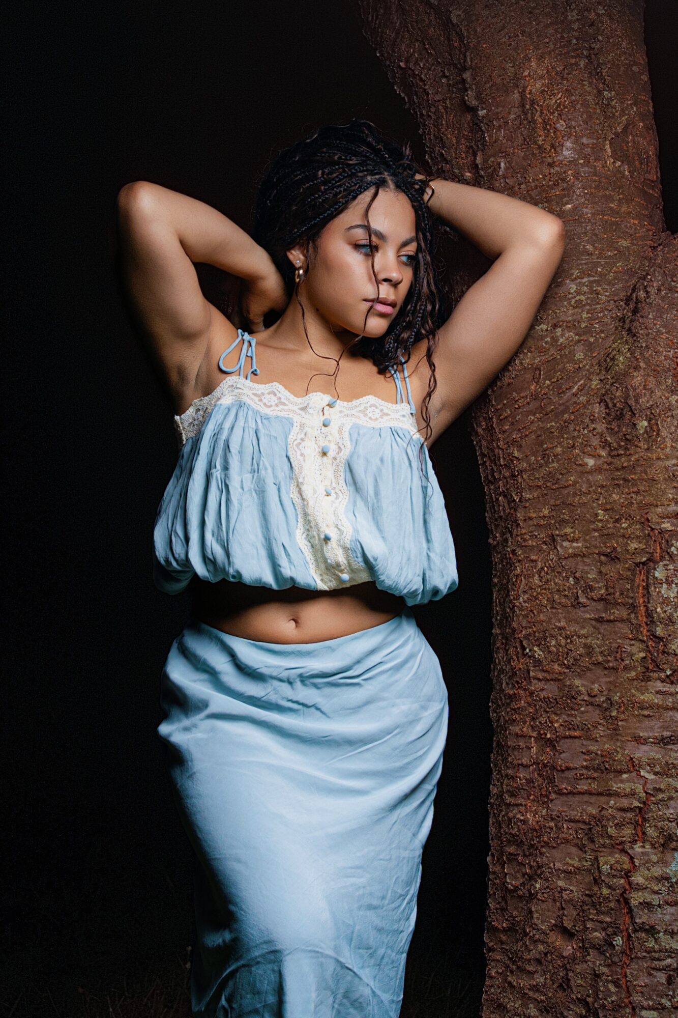 Young woman with dark hair, wearing a light blue crop top and skirt, standing against a textured wall with eyes closed.