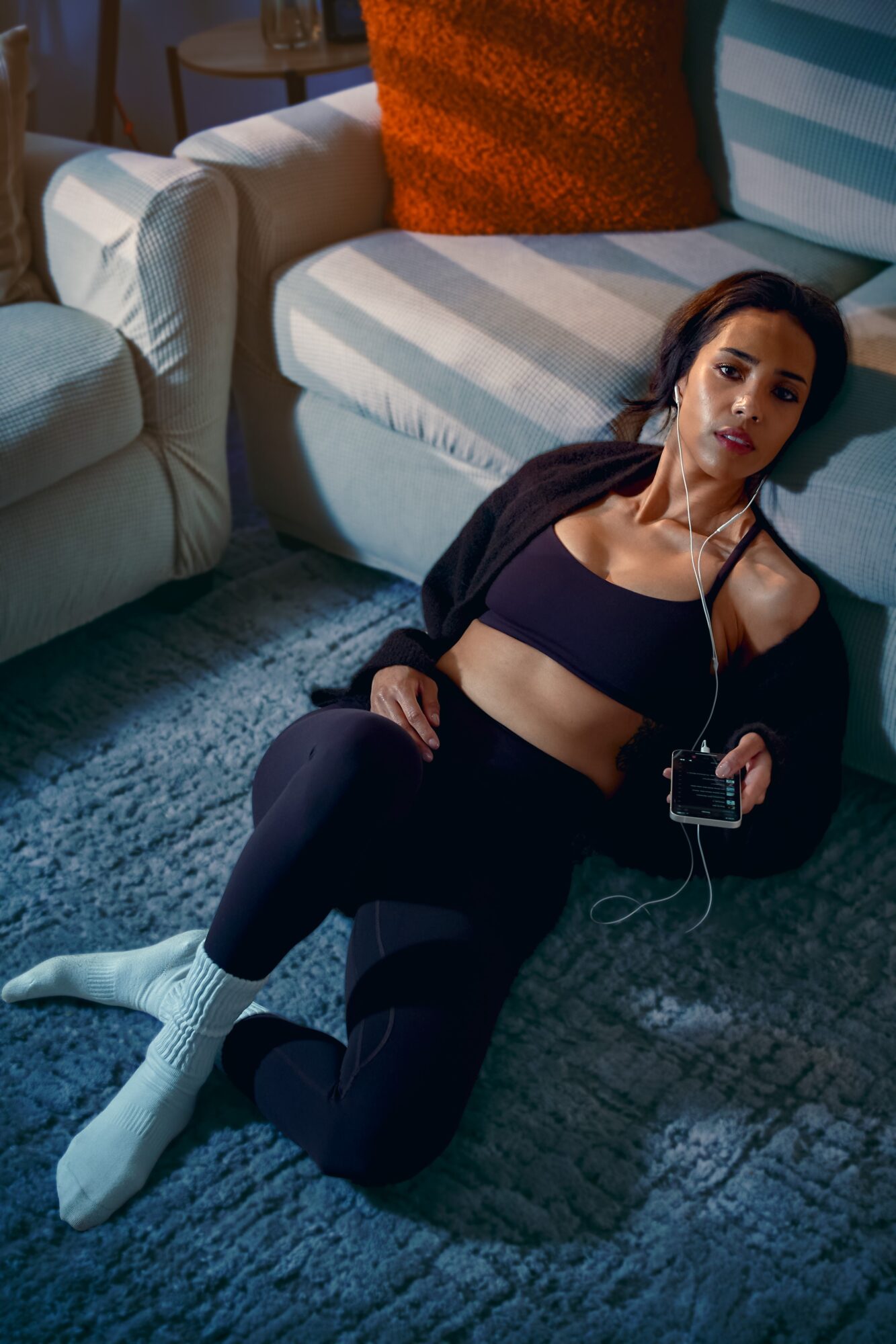Woman with long dark hair lying on the floor, wearing a black sports bra and pants, with earphones and a device, in a living room.