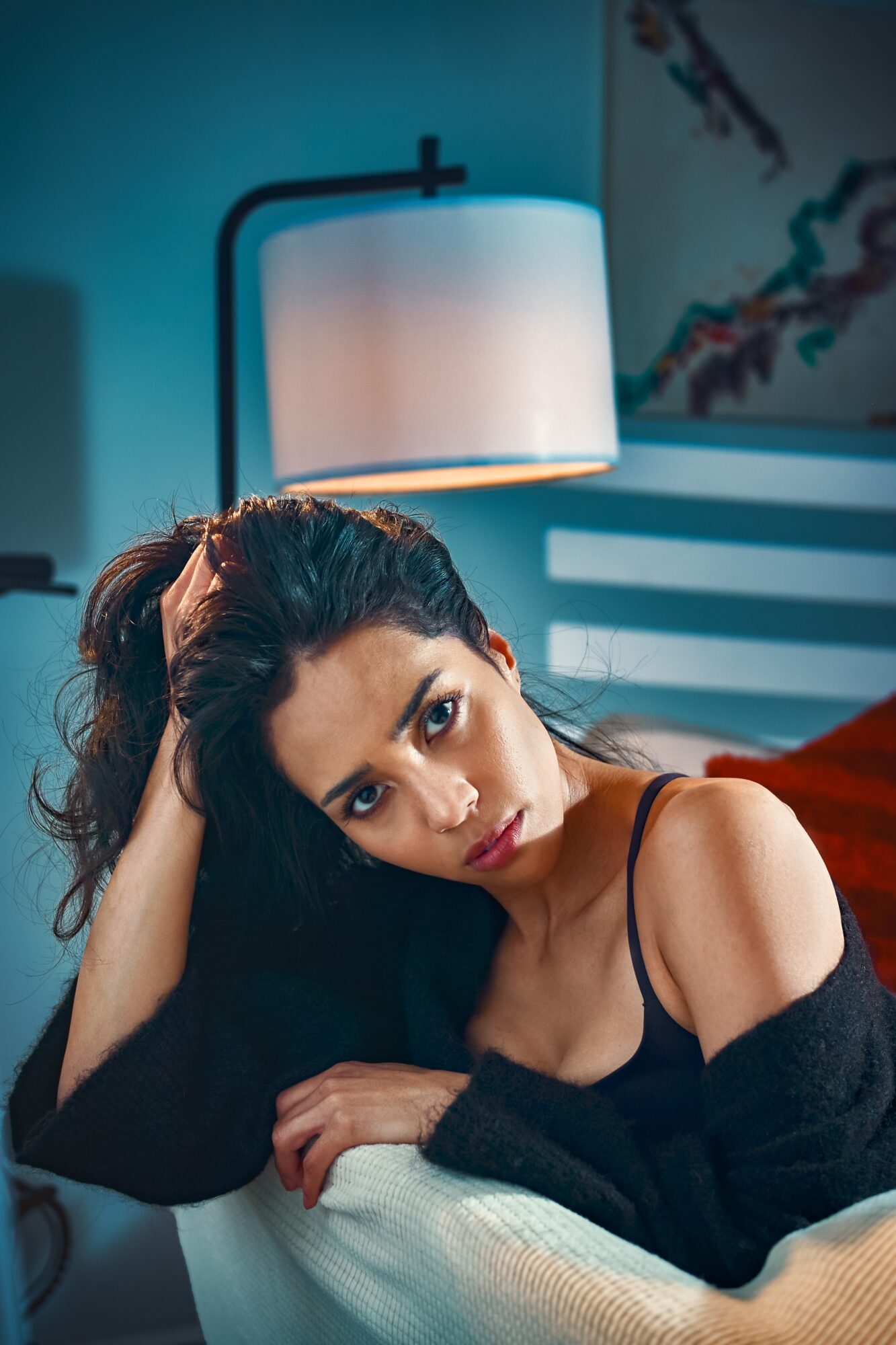 Young woman with dark hair and makeup, wearing a black top, resting her head on her hand in a dimly lit room.