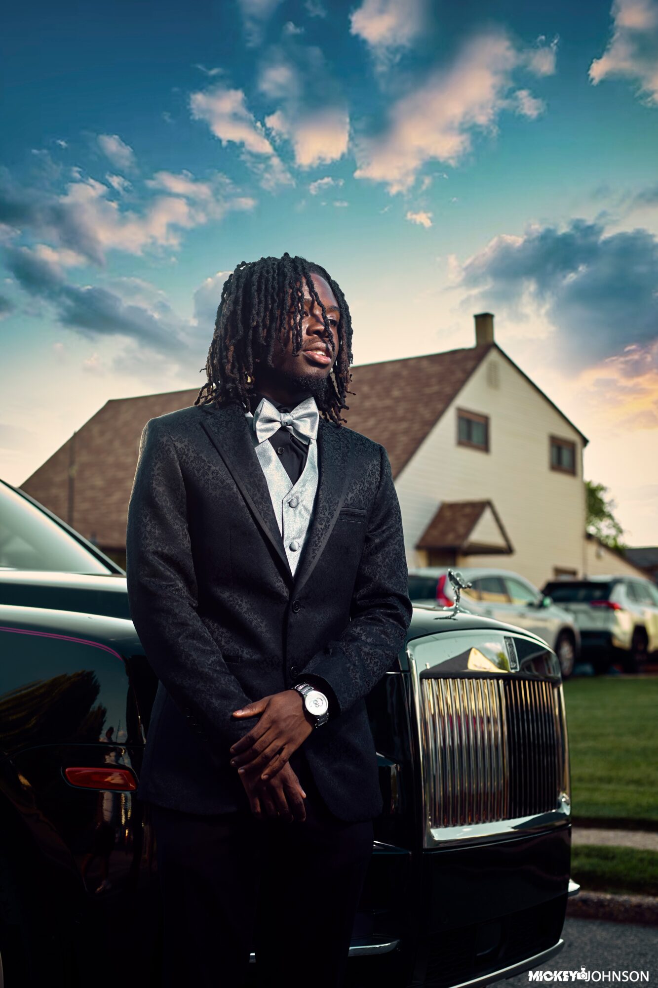 Young man in tuxedo standing outdoors near a black car, with a house and sky in the background.