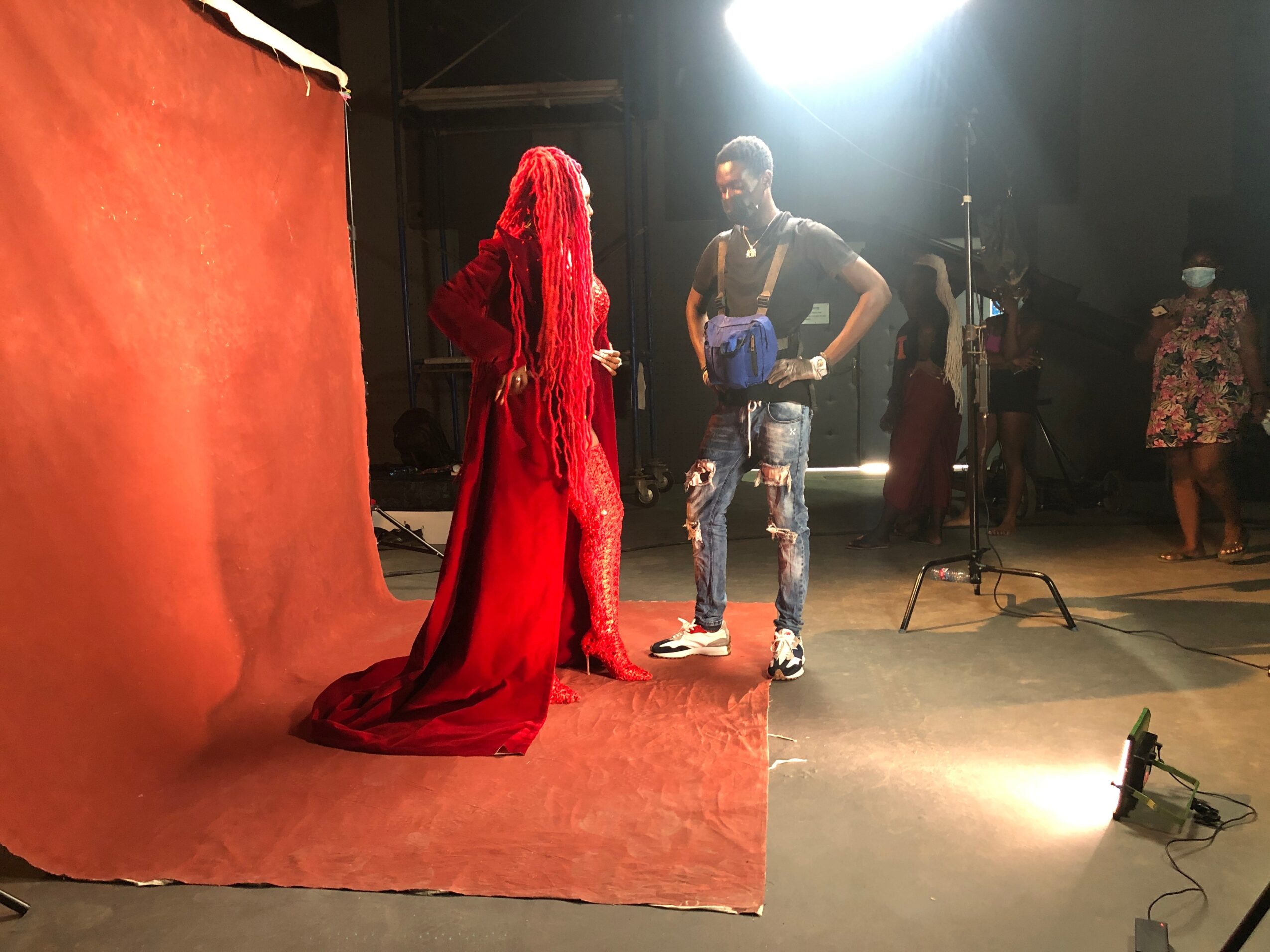 Person with long red hair and red dress facing another person in a studio setting, with others in the background.