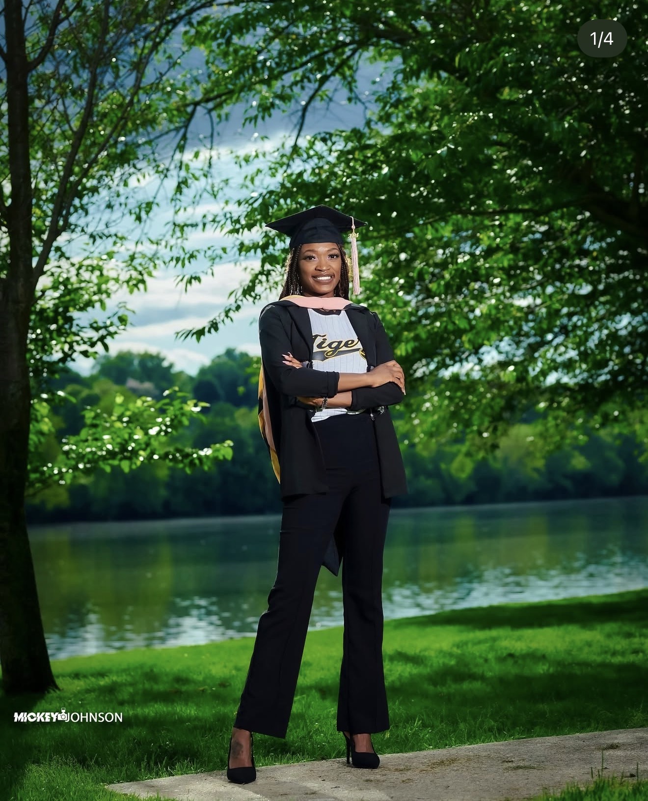 Young woman in graduation cap and gown standing outdoors by a lake with trees and sky in background.