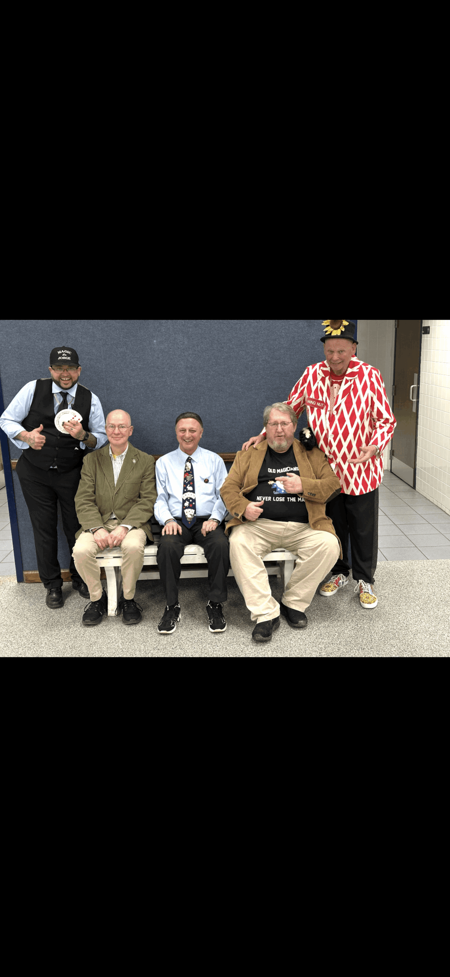 Five people sitting and standing in a room, some wearing uniforms and casual clothing, with a gray wall background.