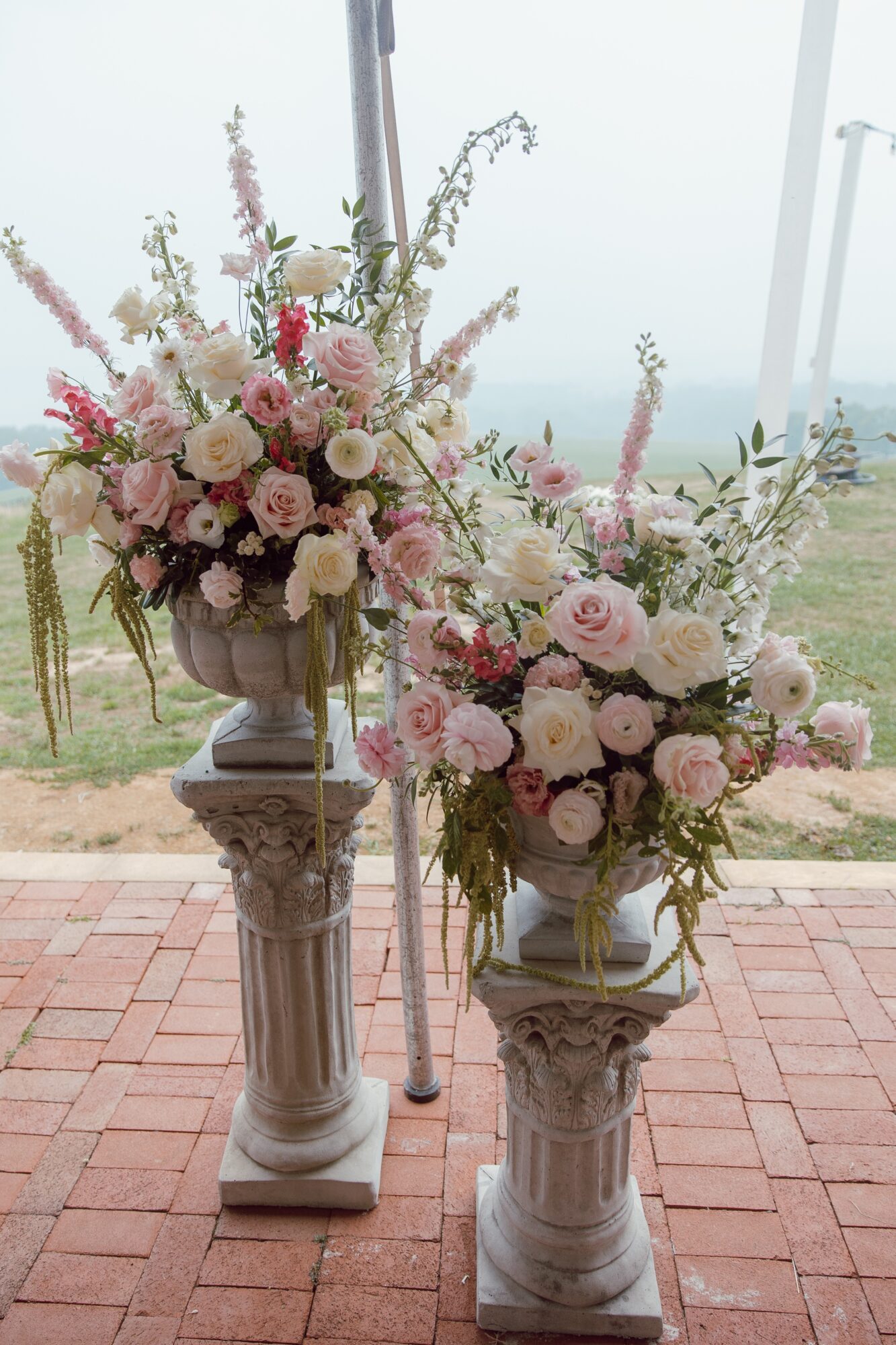 Two large floral arrangements with pink and white flowers on tall stone pedestals outdoors, with a cloudy sky and landscape in background.