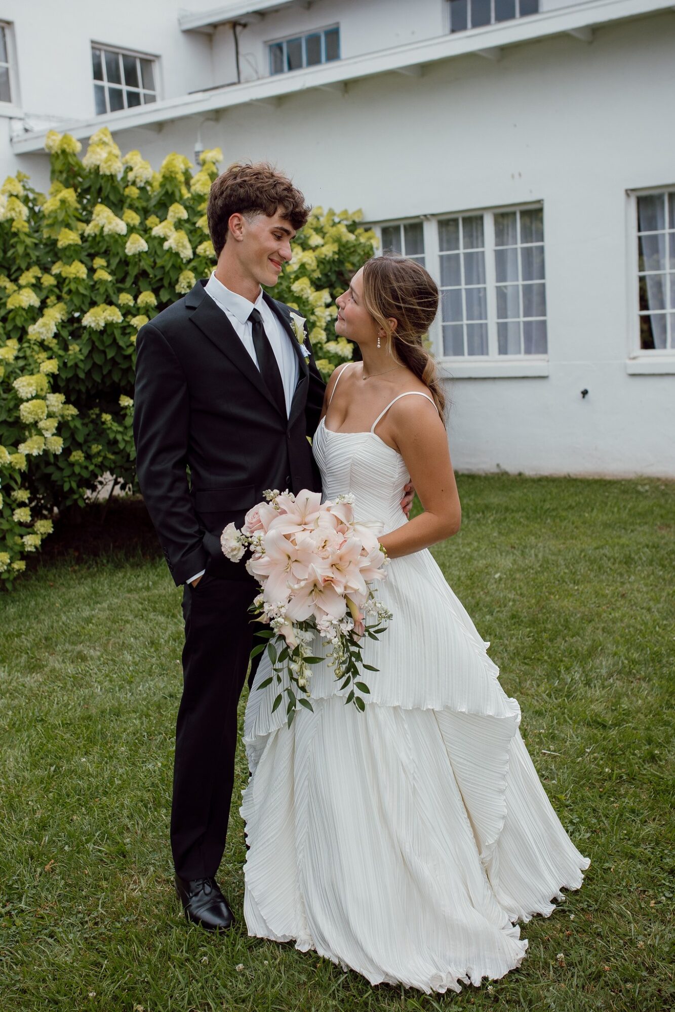 Bride and groom standing outdoors, smiling at each other, with a white building and greenery in the background.