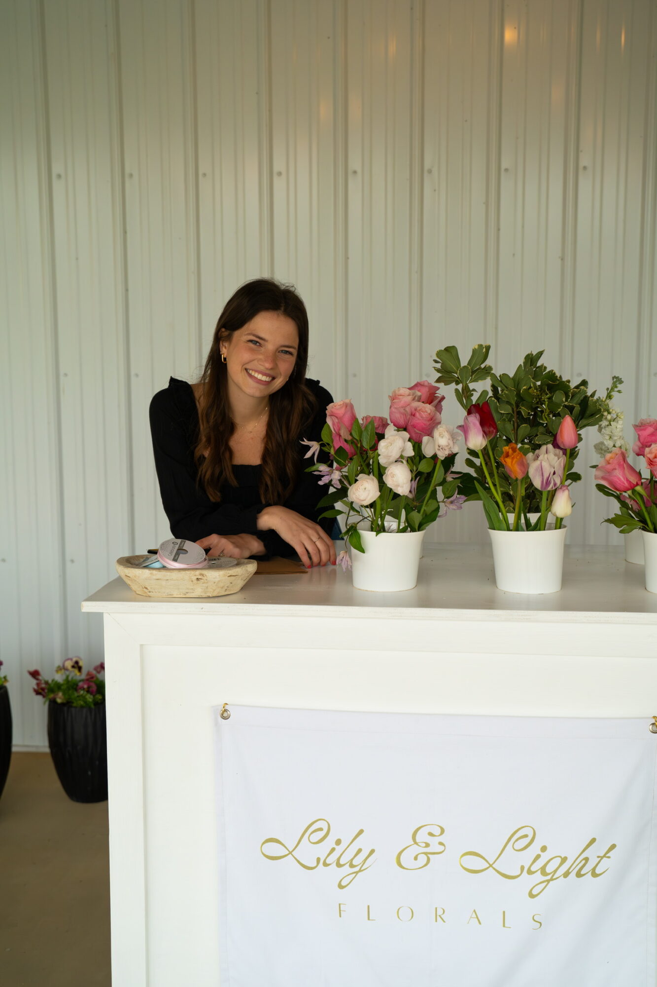 Smiling woman with long dark hair behind a white counter with pink and white flowers in vases, against a light-colored wall.