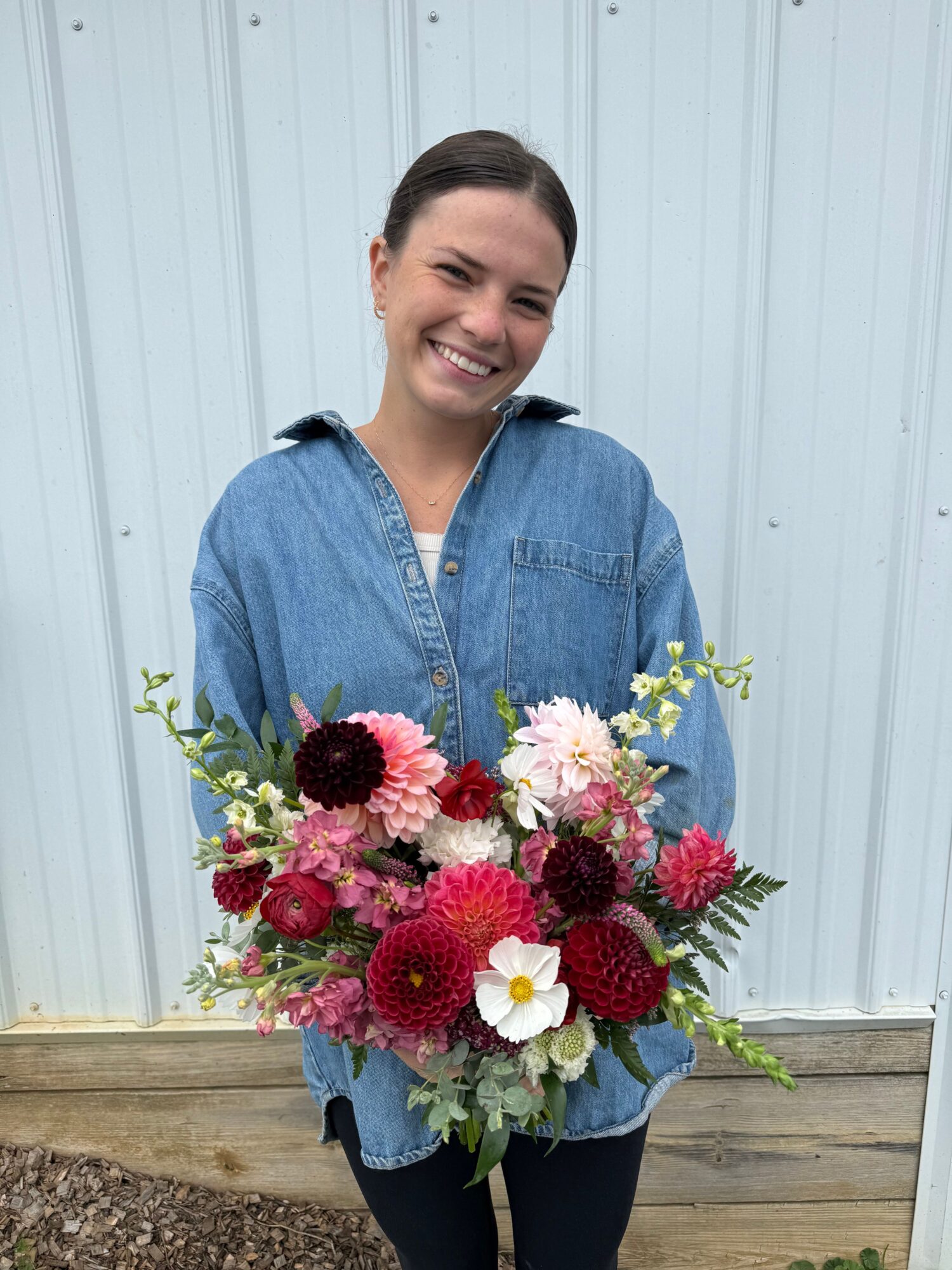 Smiling woman holding a large colorful flower bouquet, standing outdoors against a light-colored wall.