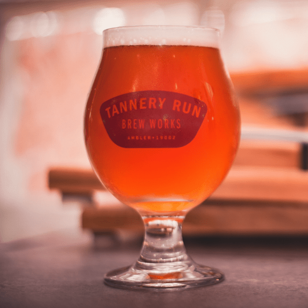 A glass of amber beer with a frothy head, labeled 'Tannery Run Brew Works,' on a dark surface with blurred background.