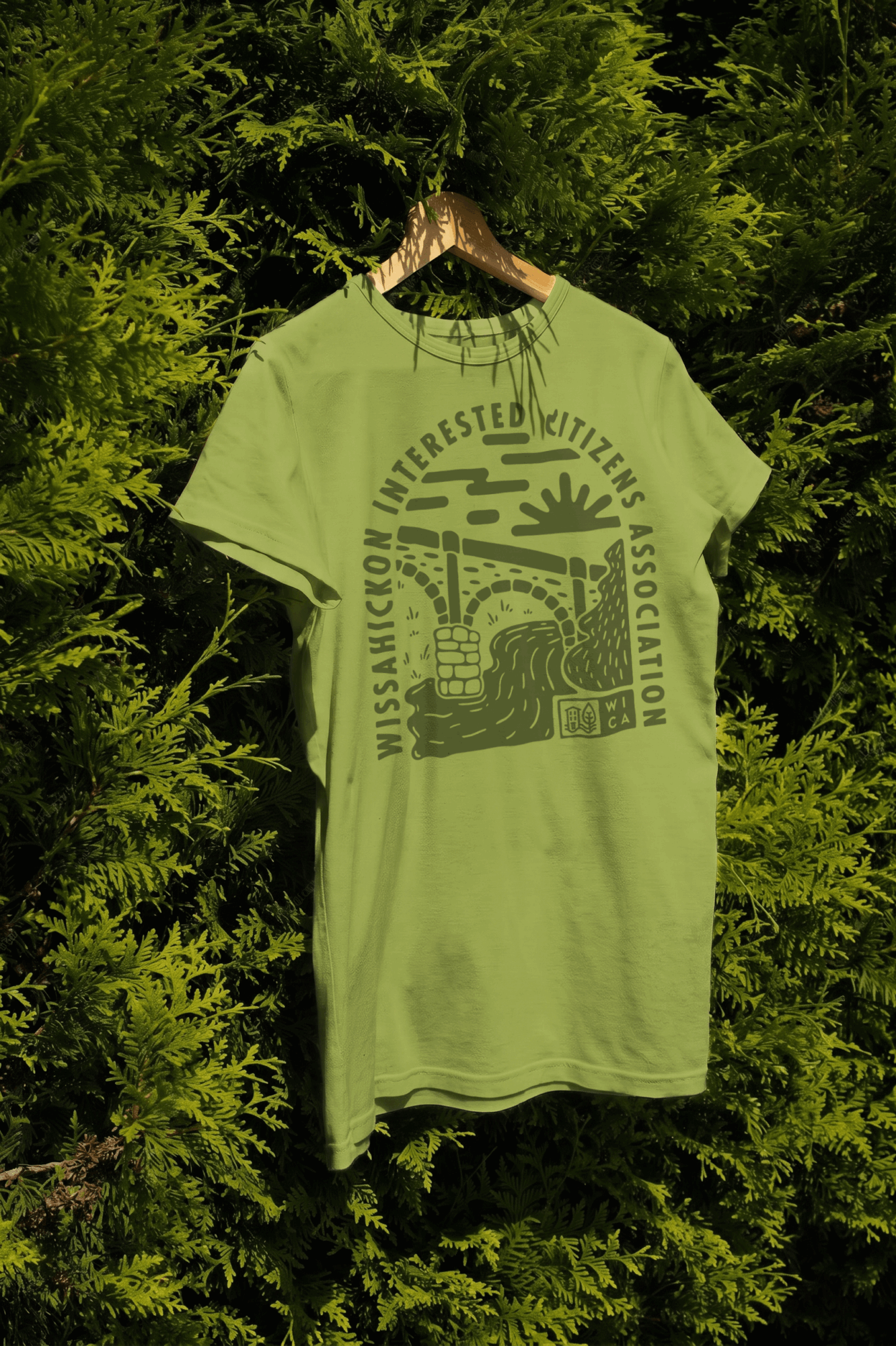 Light green T-shirt with a graphic of a crocodile and text, hanging on a hanger among green foliage.
