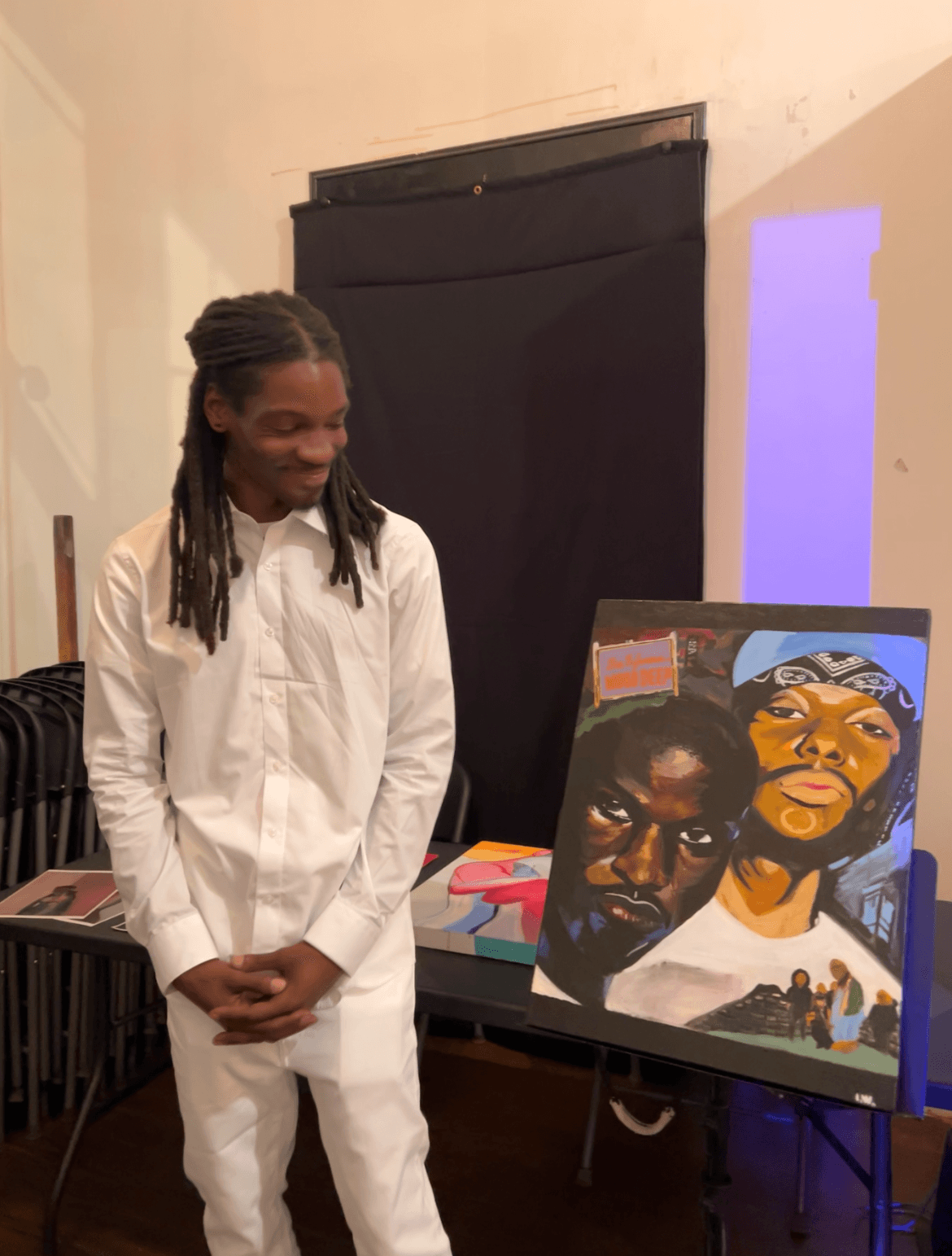 Young girl with braided hair wearing a white outfit, standing indoors near a colorful painting on an easel.