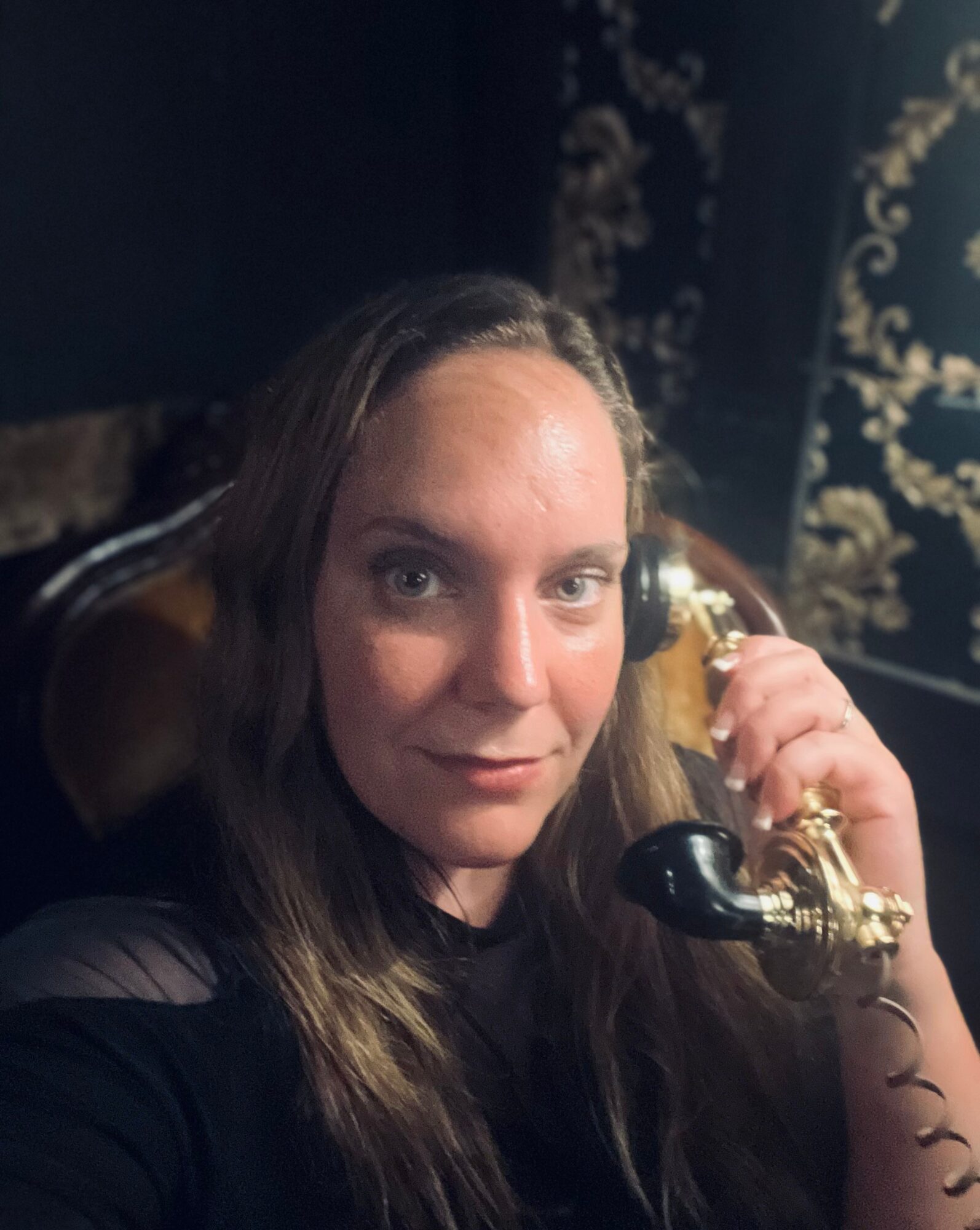 Woman with long hair holding a vintage black and gold telephone to her ear, smiling slightly, dark background with ornate patterns