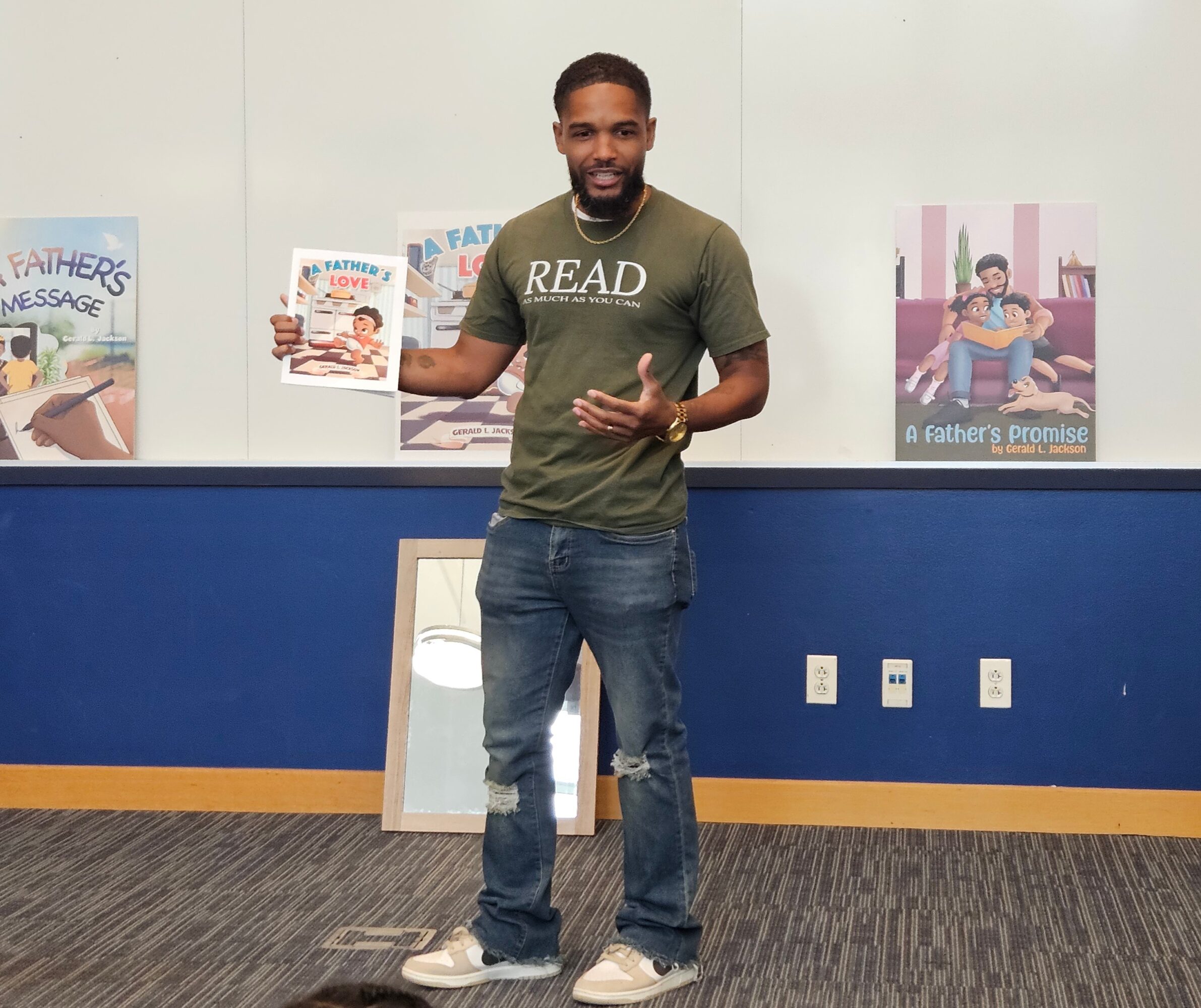 Man standing in front of a wall with children's books and posters, holding a book in his right hand, smiling.