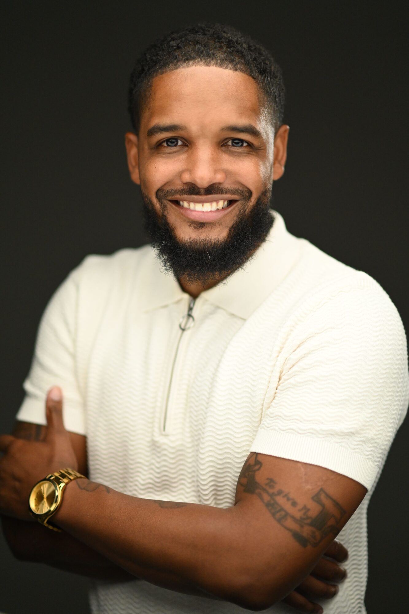 Smiling man with a beard and tattoos, wearing a white shirt, against a dark background.