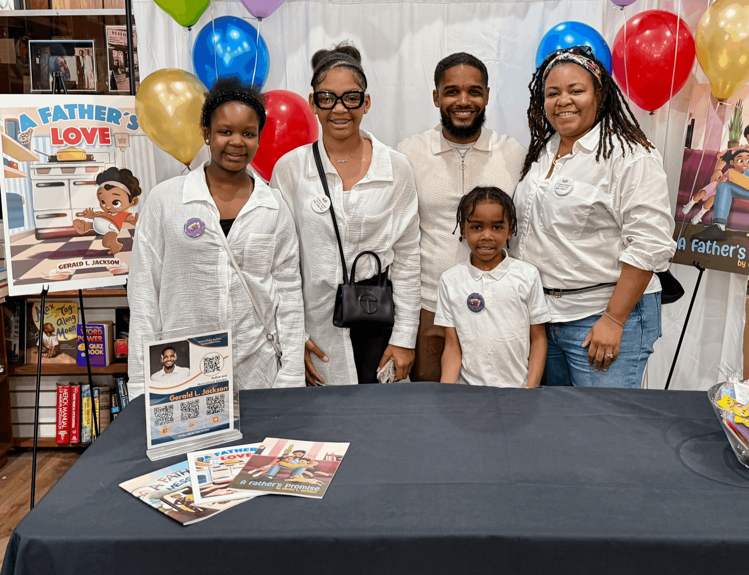 Group of five people, including children, standing behind a table with flyers, in front of colorful balloons and books.