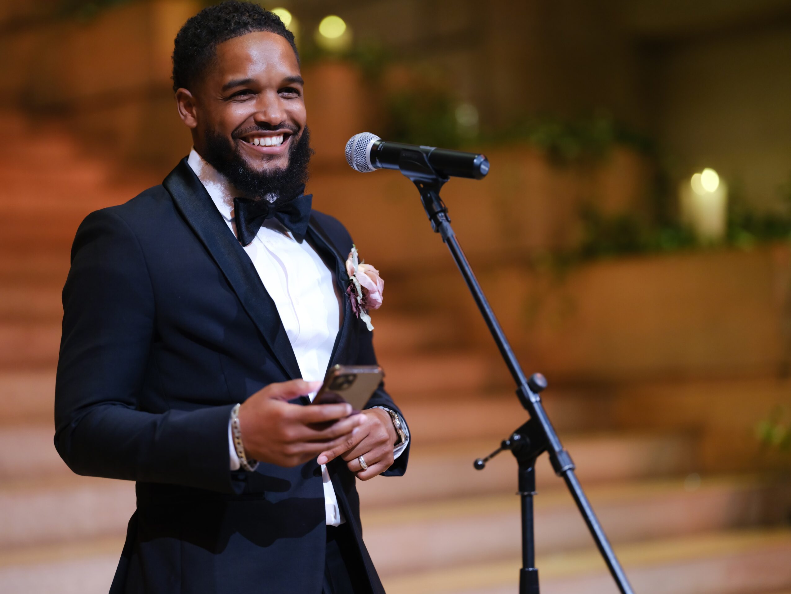 Man in tuxedo smiling at microphone, holding a phone, in an indoor setting with warm lighting and plants.