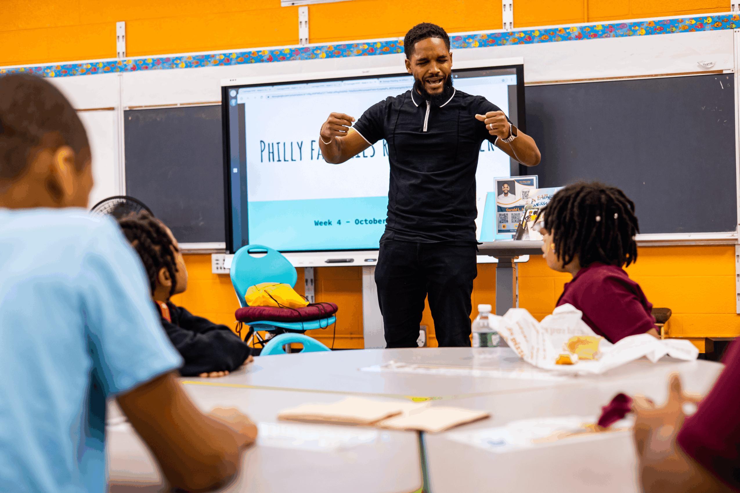 Teacher standing in front of students in classroom, gesturing with arms, blackboard behind, students seated at desks, classroom setting.