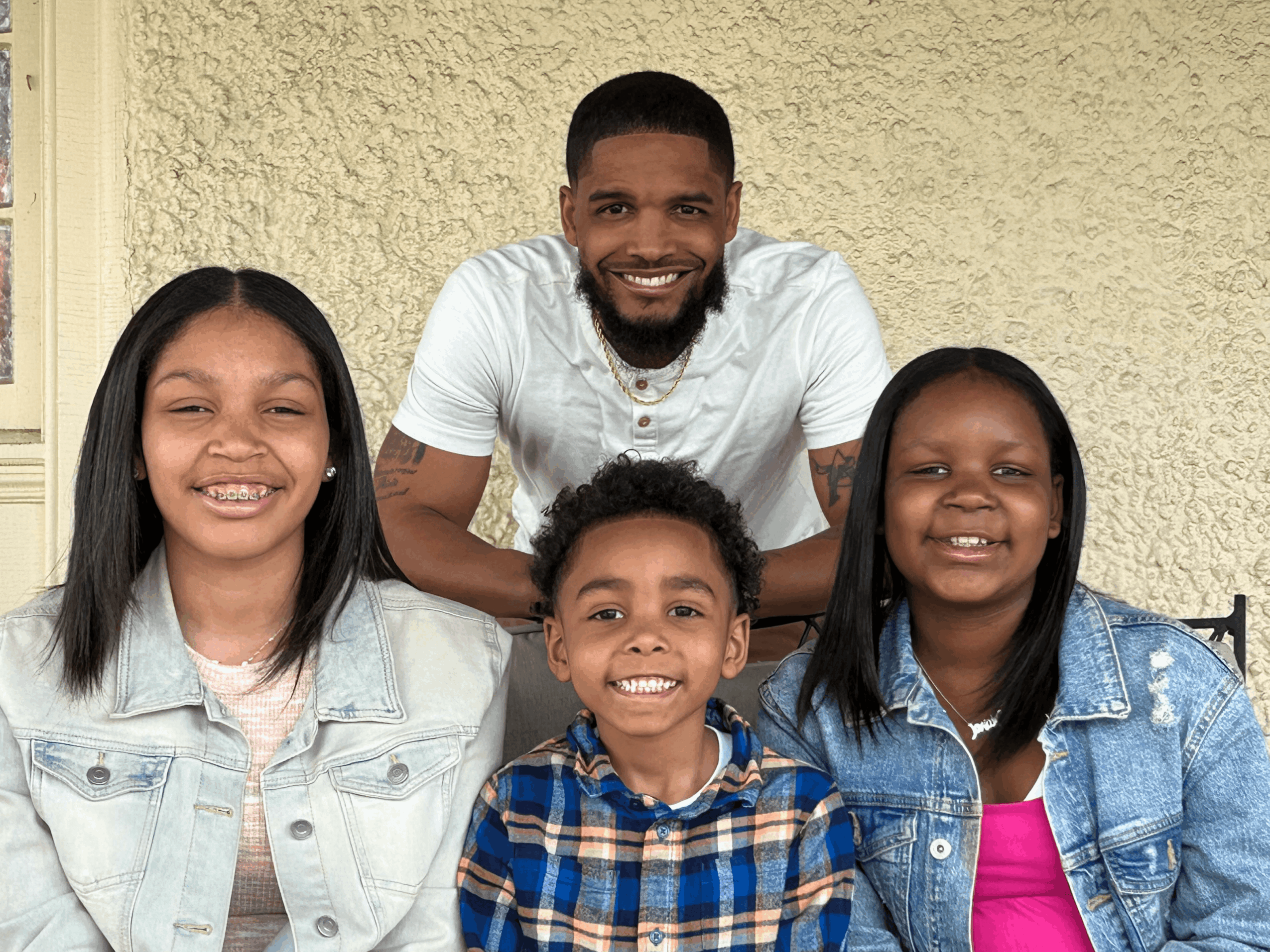 Four people smiling, three children and one adult, standing outdoors against a beige wall.