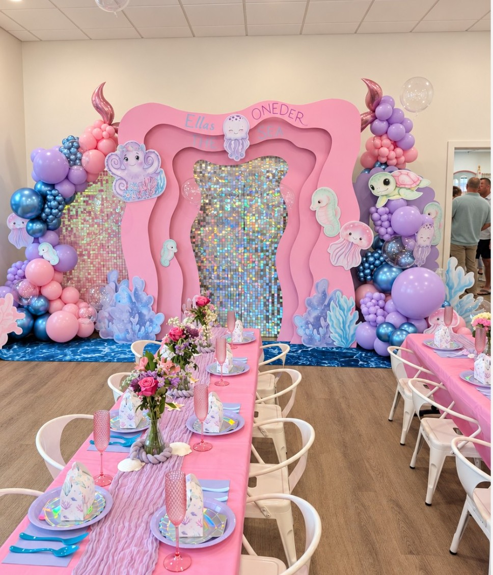 Decorative pink and purple balloon arch with unicorn and cloud motifs, backdrop with colorful mosaic, set for a celebration.