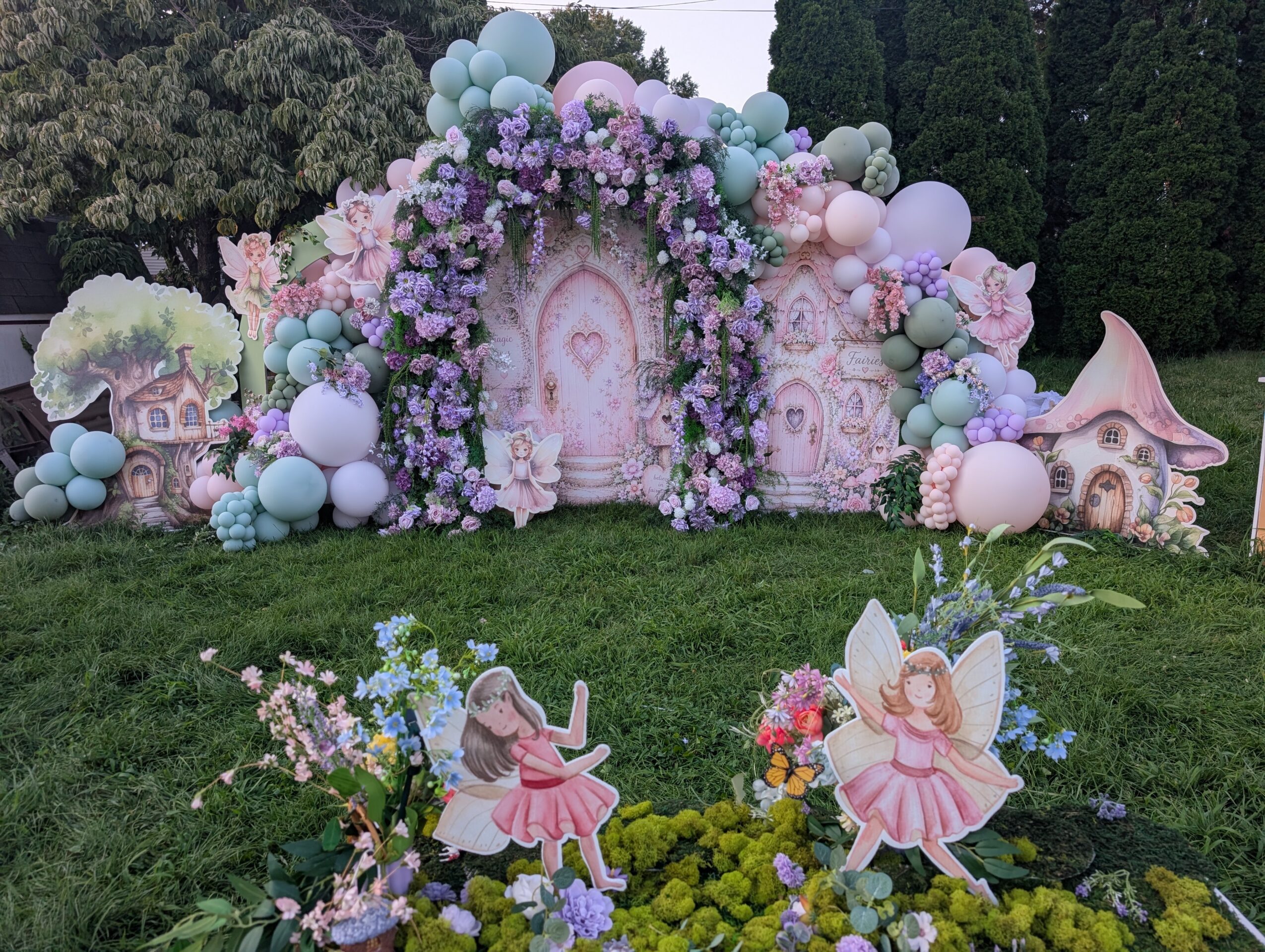 Decorative fairy tale scene with pastel-colored balloons, flowers, and fairy cutouts outdoors on grass.