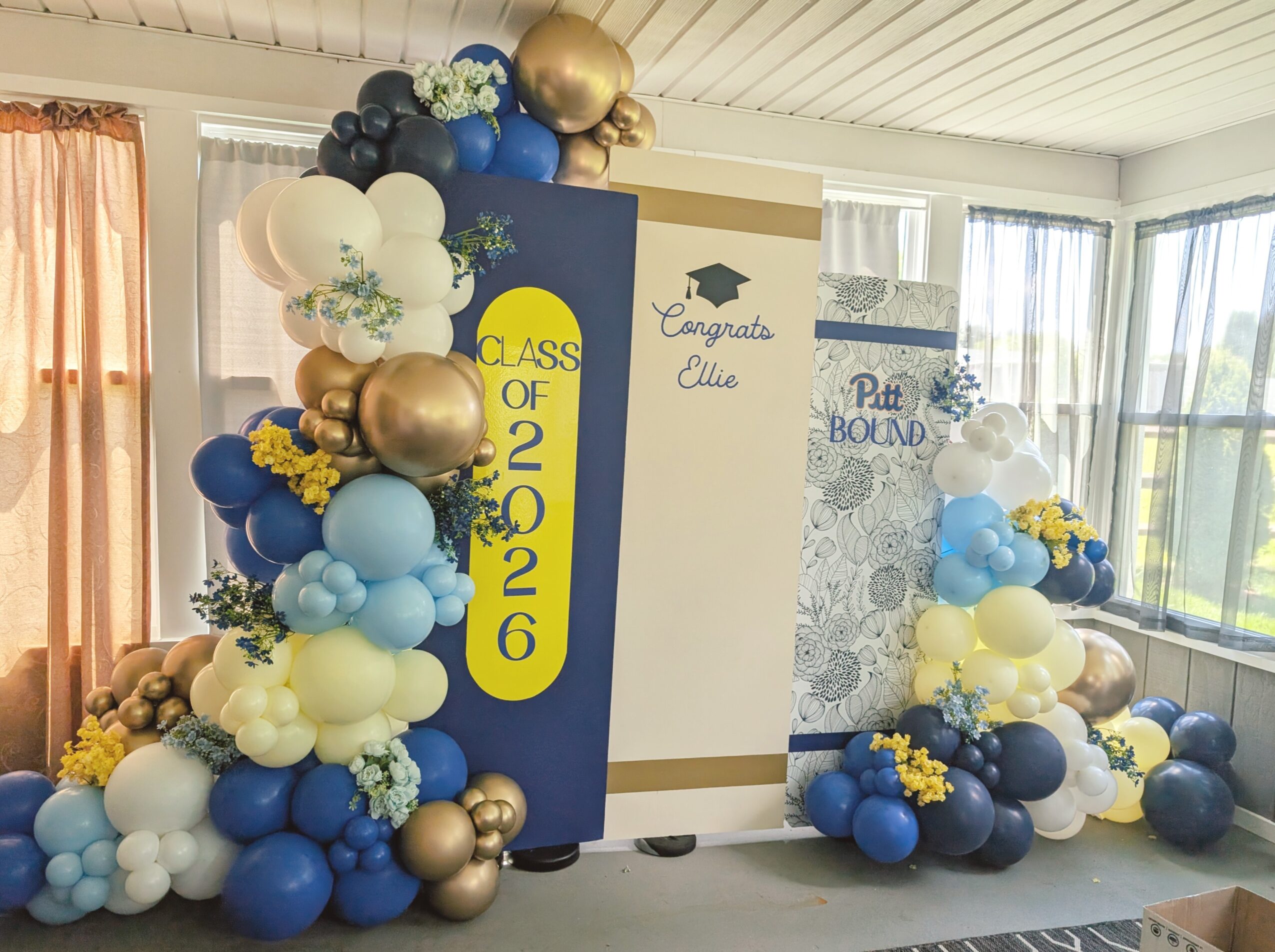 Decorative graduation display with blue, white, gold, and black balloons, a congratulatory sign, and a backdrop with a graduation cap.