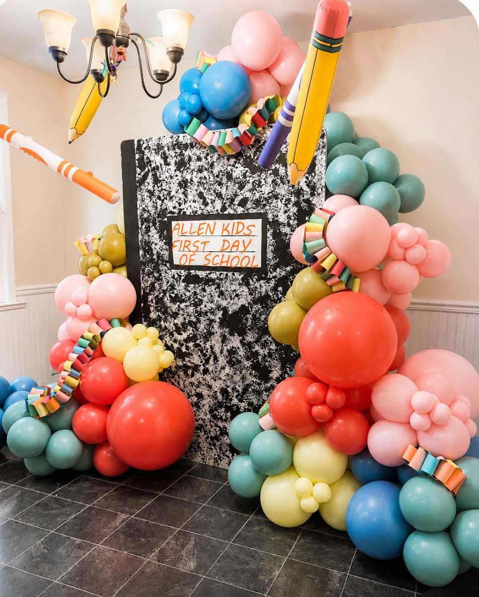 Decorative display with colorful balloons, a black and white speckled board, and a sign reading 'Fallen Kids First Day of School' in a room with a chandelier and tiled floor.