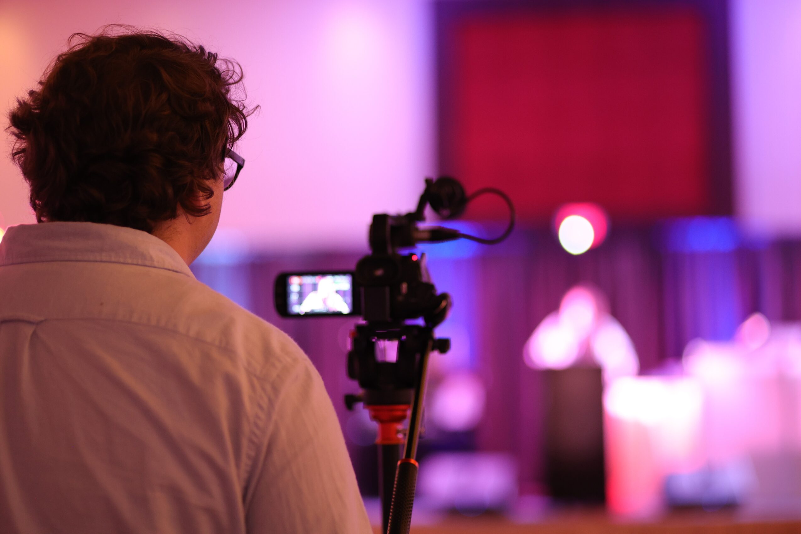 Person filming a stage with a camera, blurred background with stage lights and seating area.