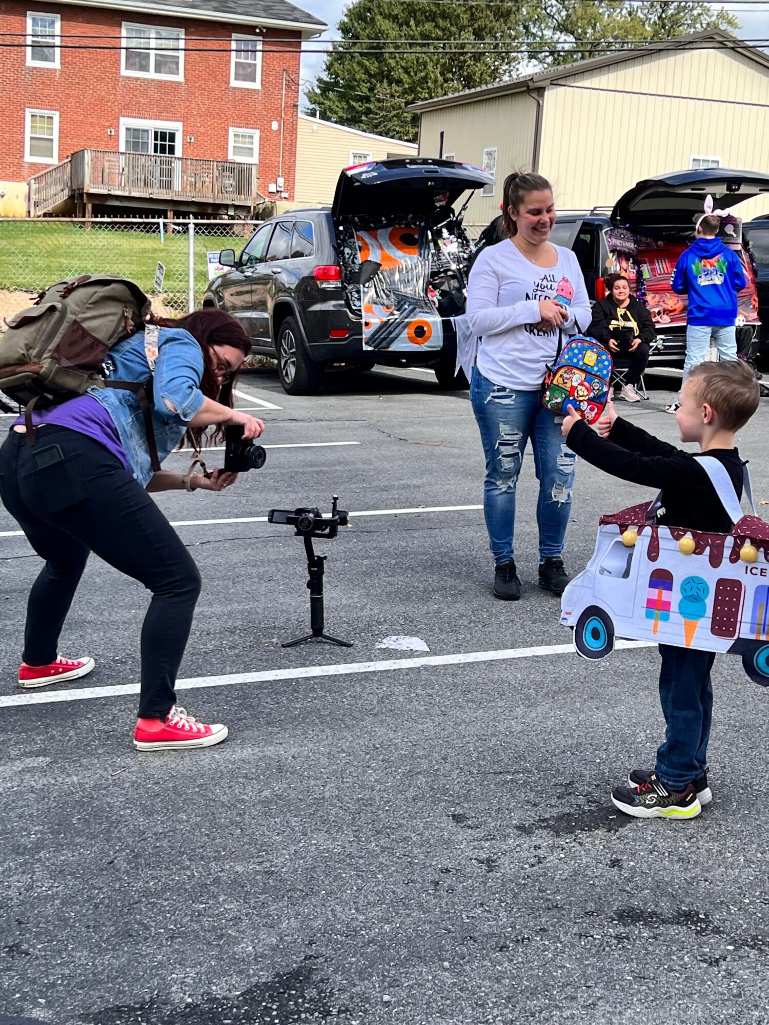 People gather around a table with toys and items outdoors, with a woman taking a photo and children present.