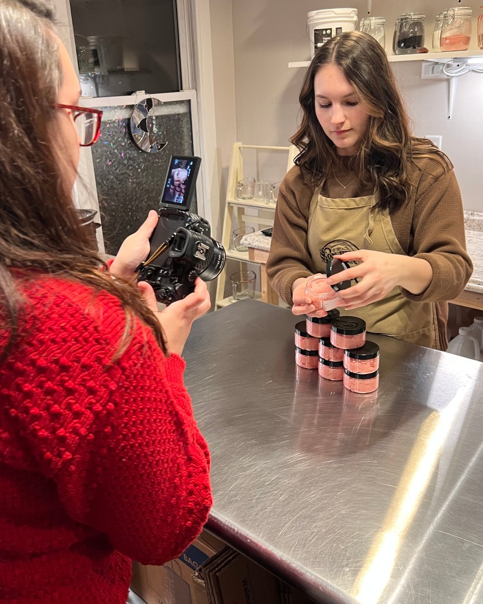 Two women in a kitchen, one taking a photo of the other stacking small jars on a table, with shelves and containers in the background.