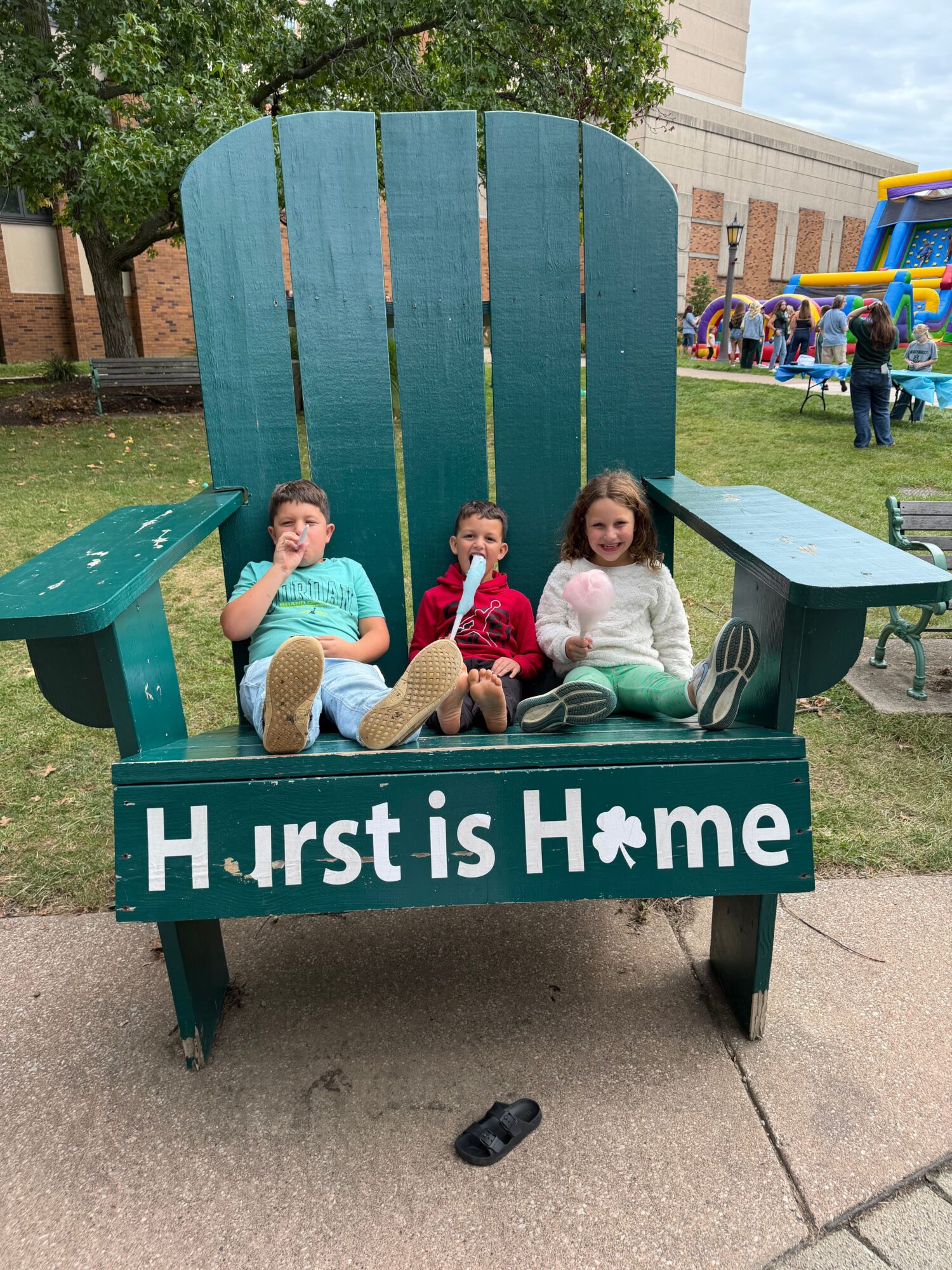Three children sit behind a sign that reads 'Hust is Hame' at a playground, with colorful equipment in the background.