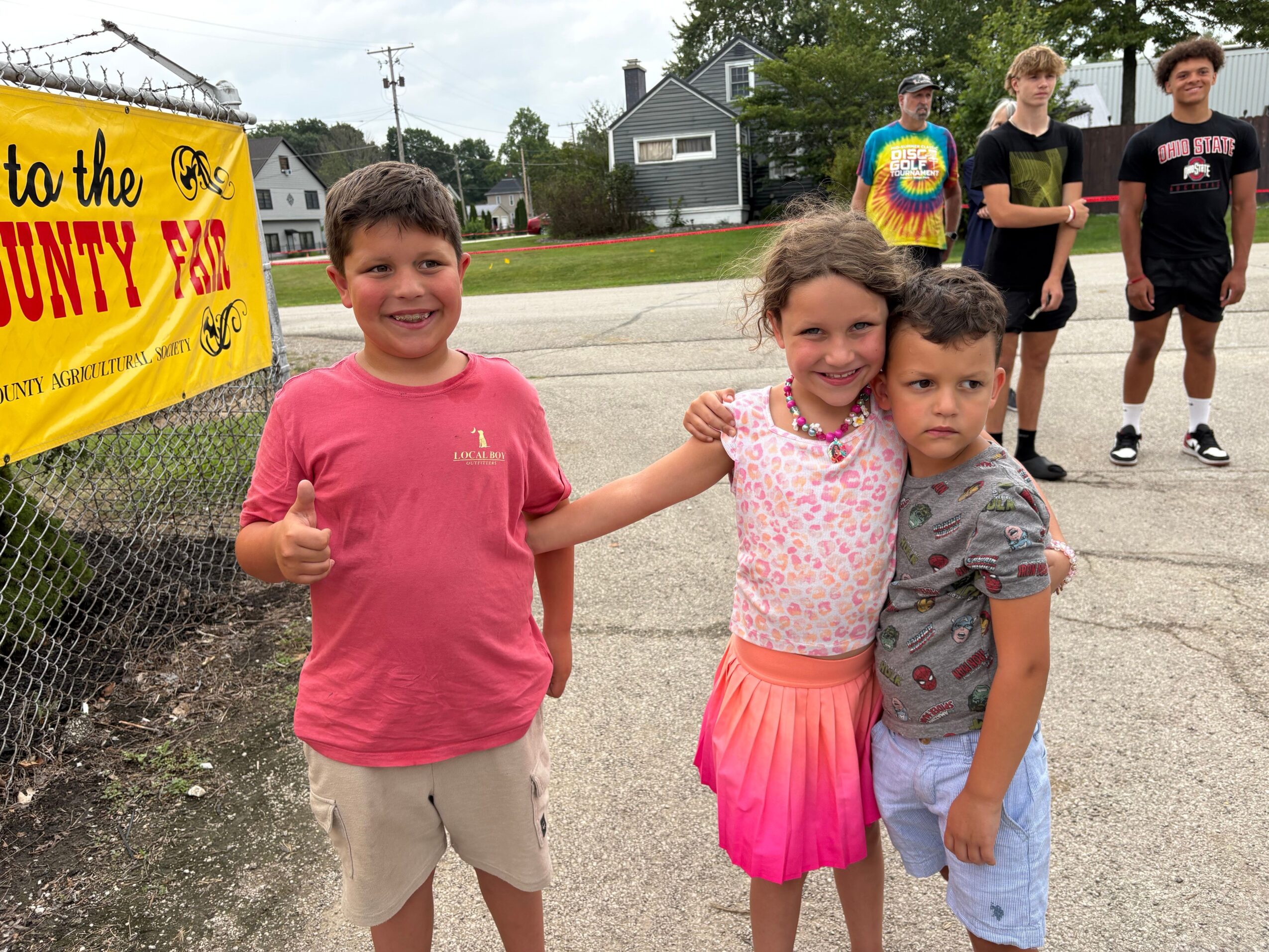 Four children standing outdoors, two girls in pink dresses and two boys in casual clothes, with a yellow sign in the background.