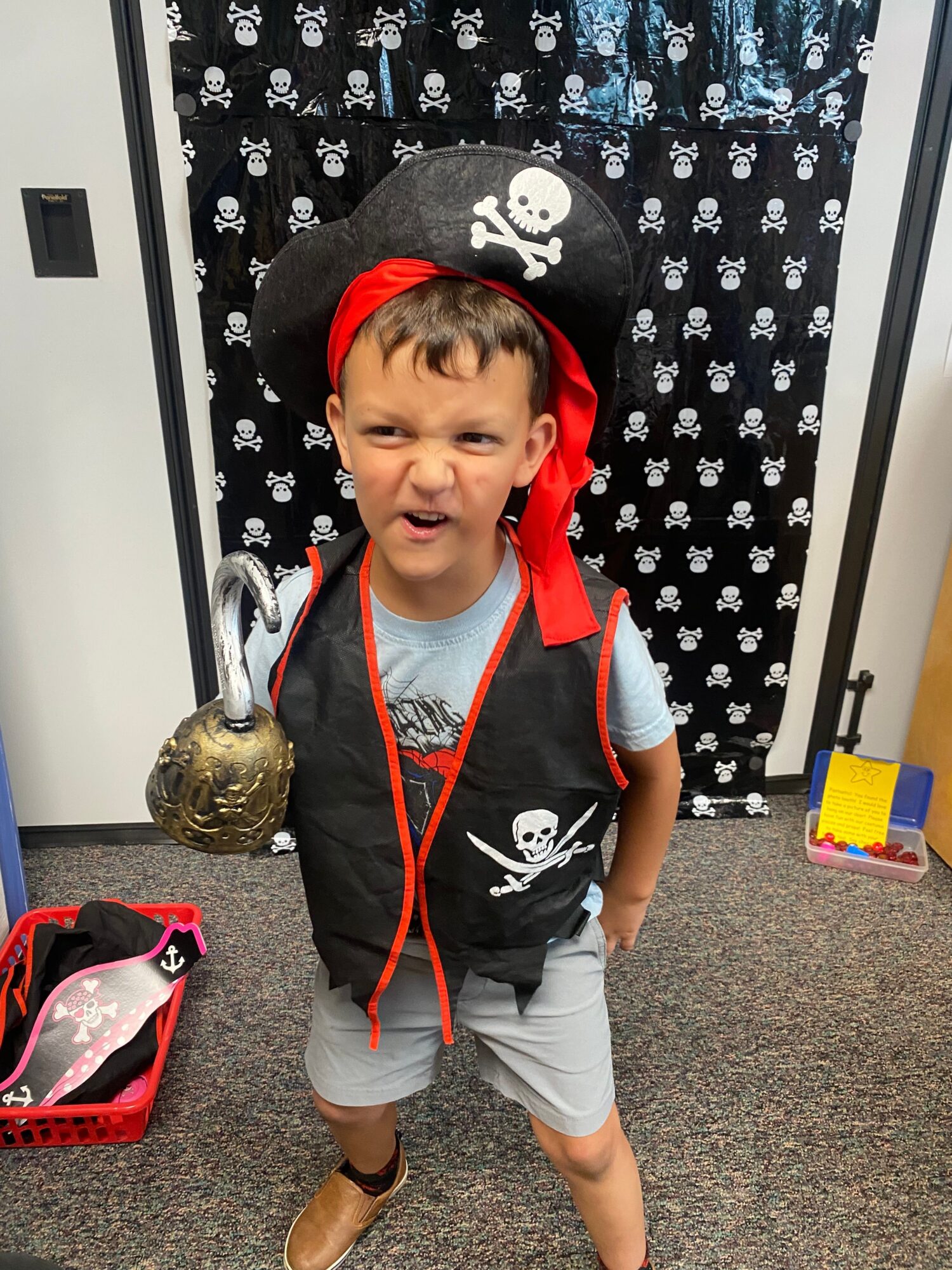 Child dressed as a pirate with a hat, standing indoors near a black and white curtain, holding a gold-colored object, with a pink bag nearby.